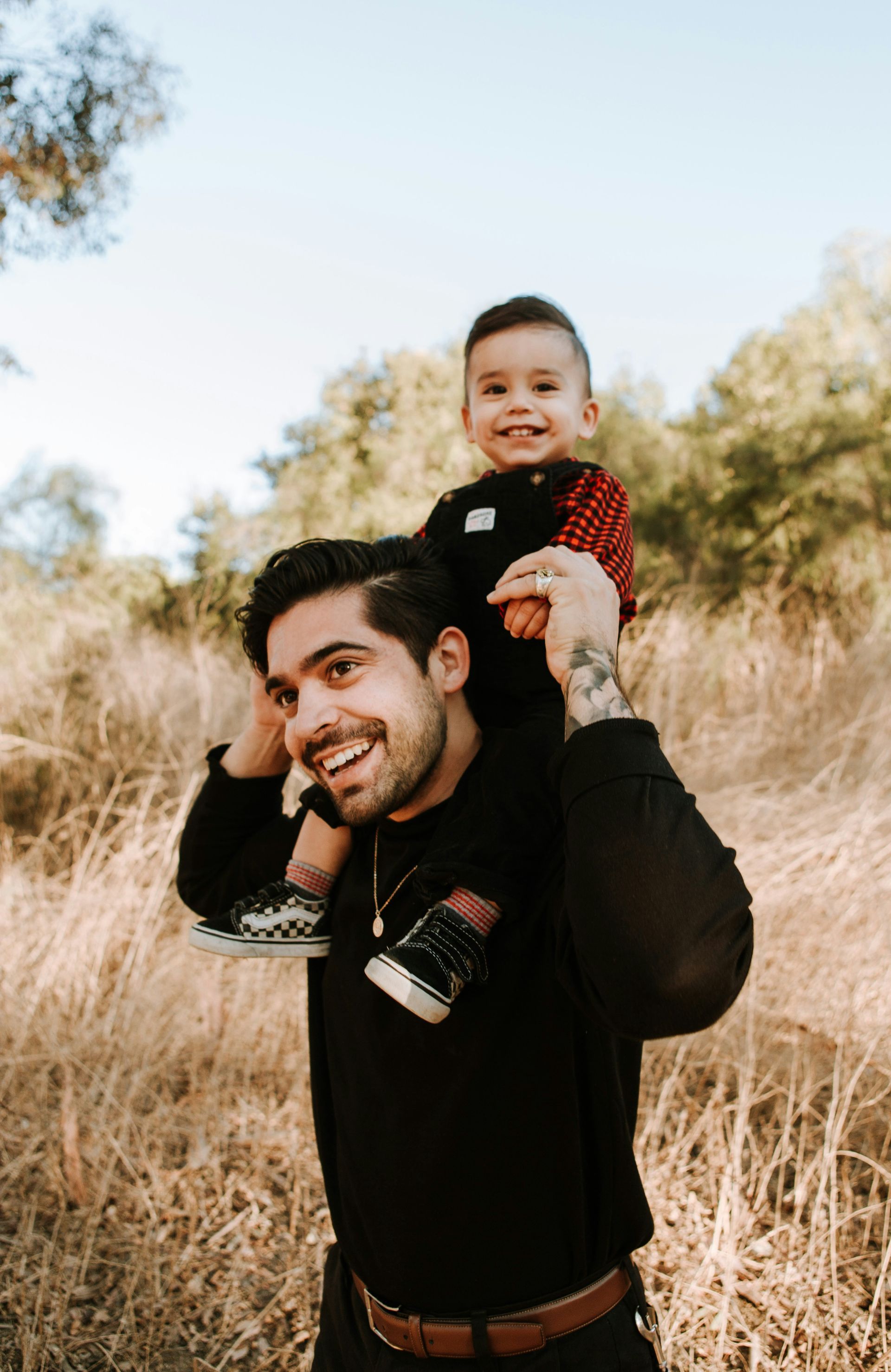 Man carrying a smiling child on his shoulders in a field. The man smiles, the child wears a red shirt.