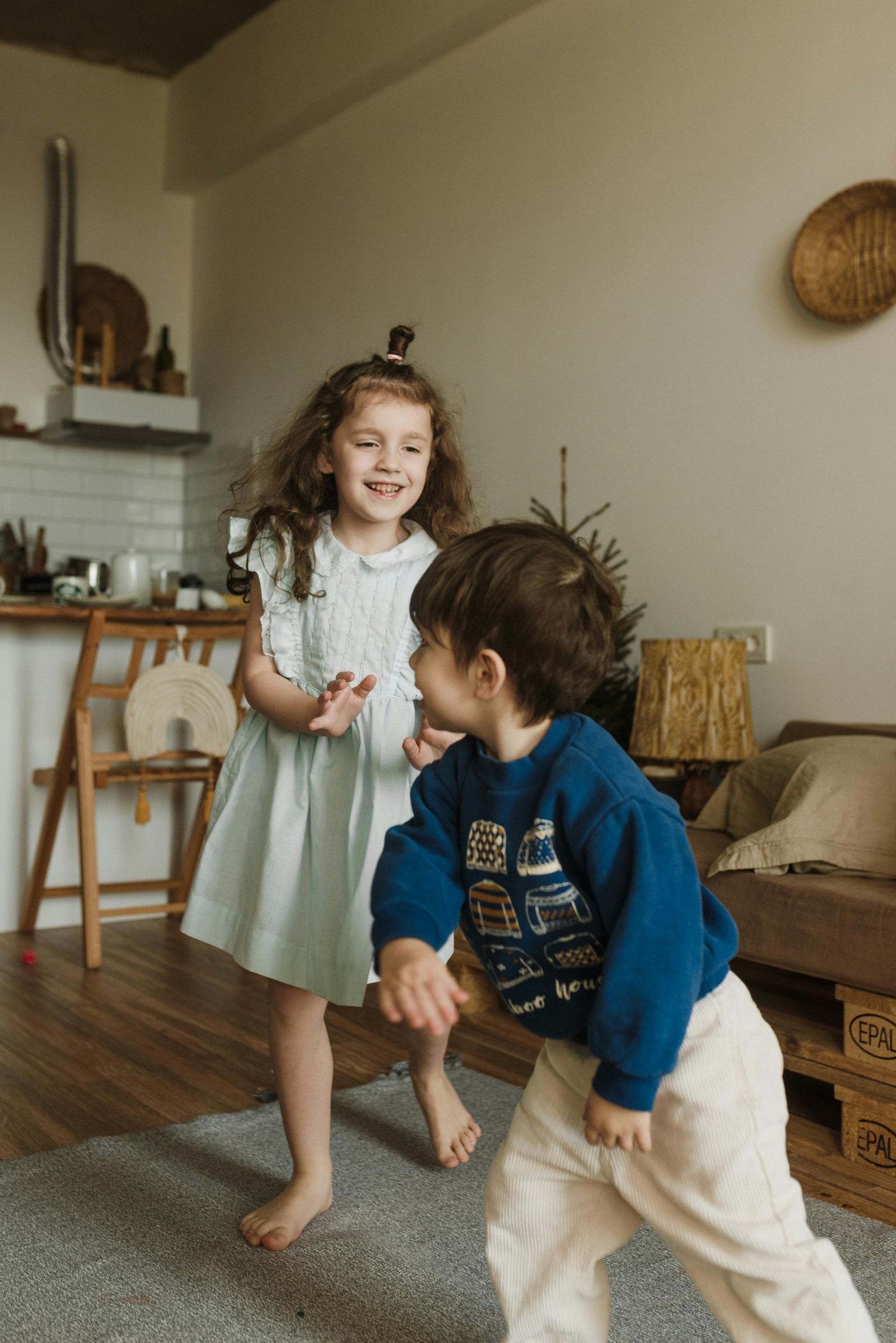 Girl and boy laughing, dancing in a living room. Girl wears blue dress, boy blue sweater.