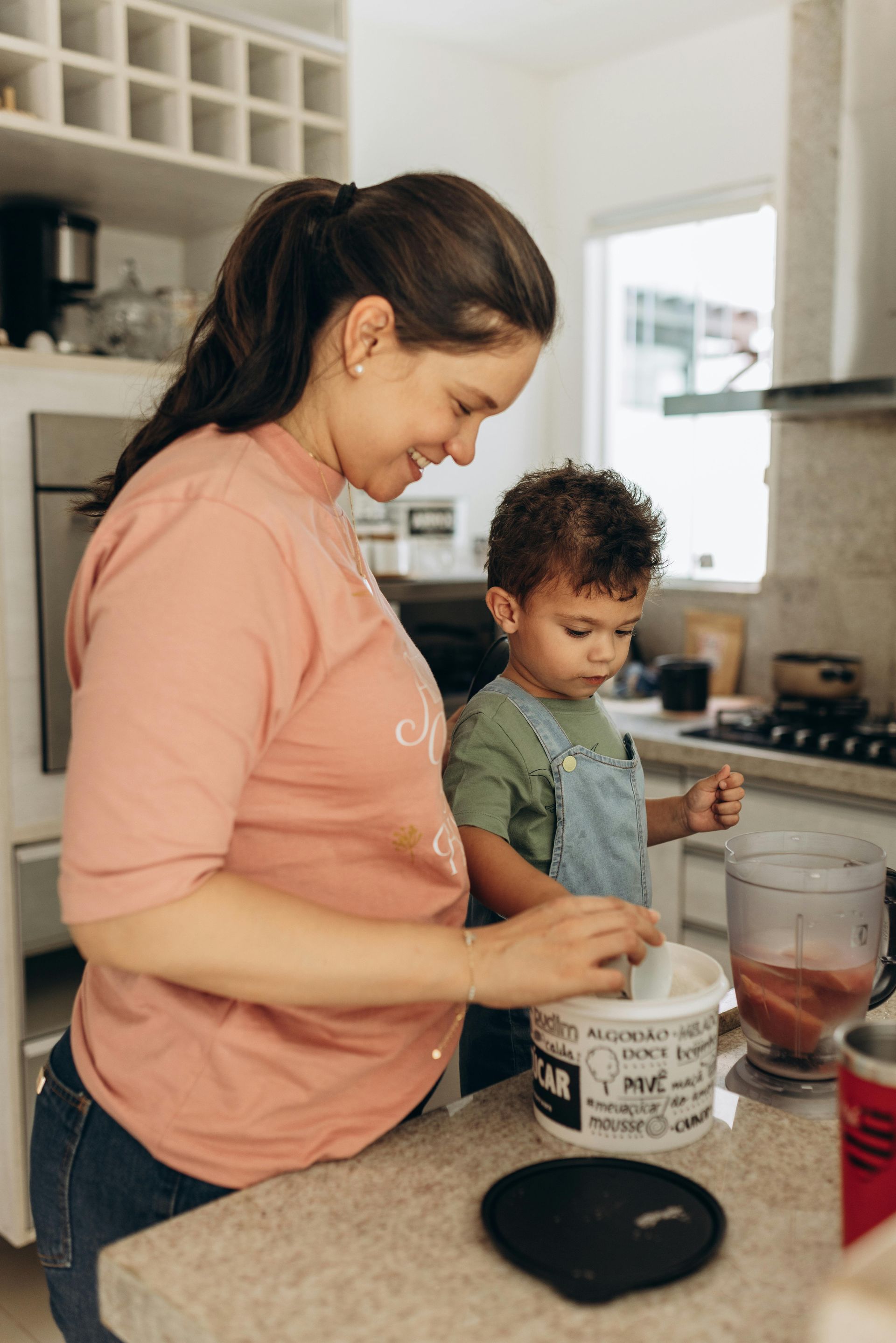 Woman and child in kitchen, making something, child wearing overalls, woman pink shirt, smiling.