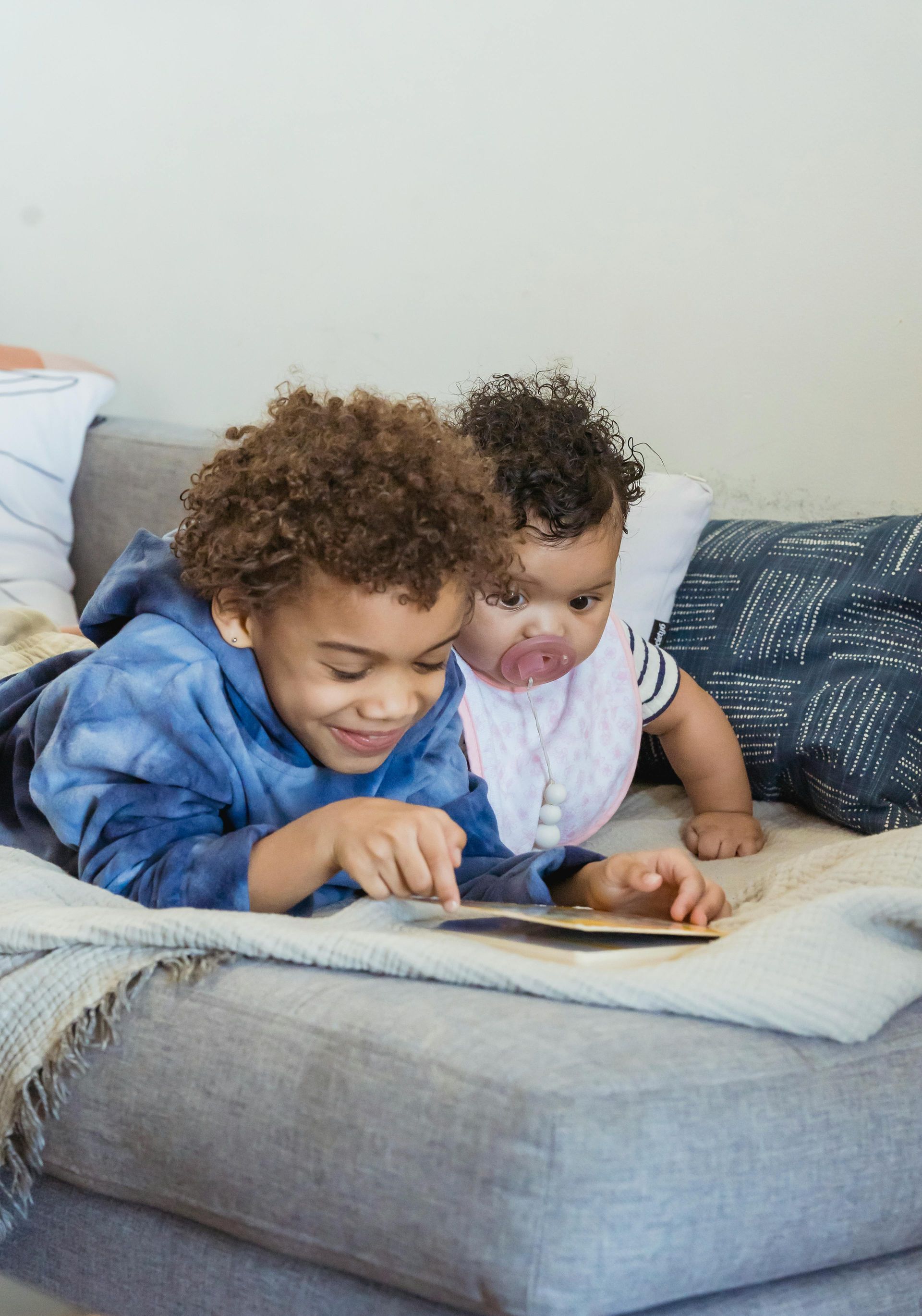 Two children, one older, reading a book together on a gray couch. The baby wears a bib and watches.