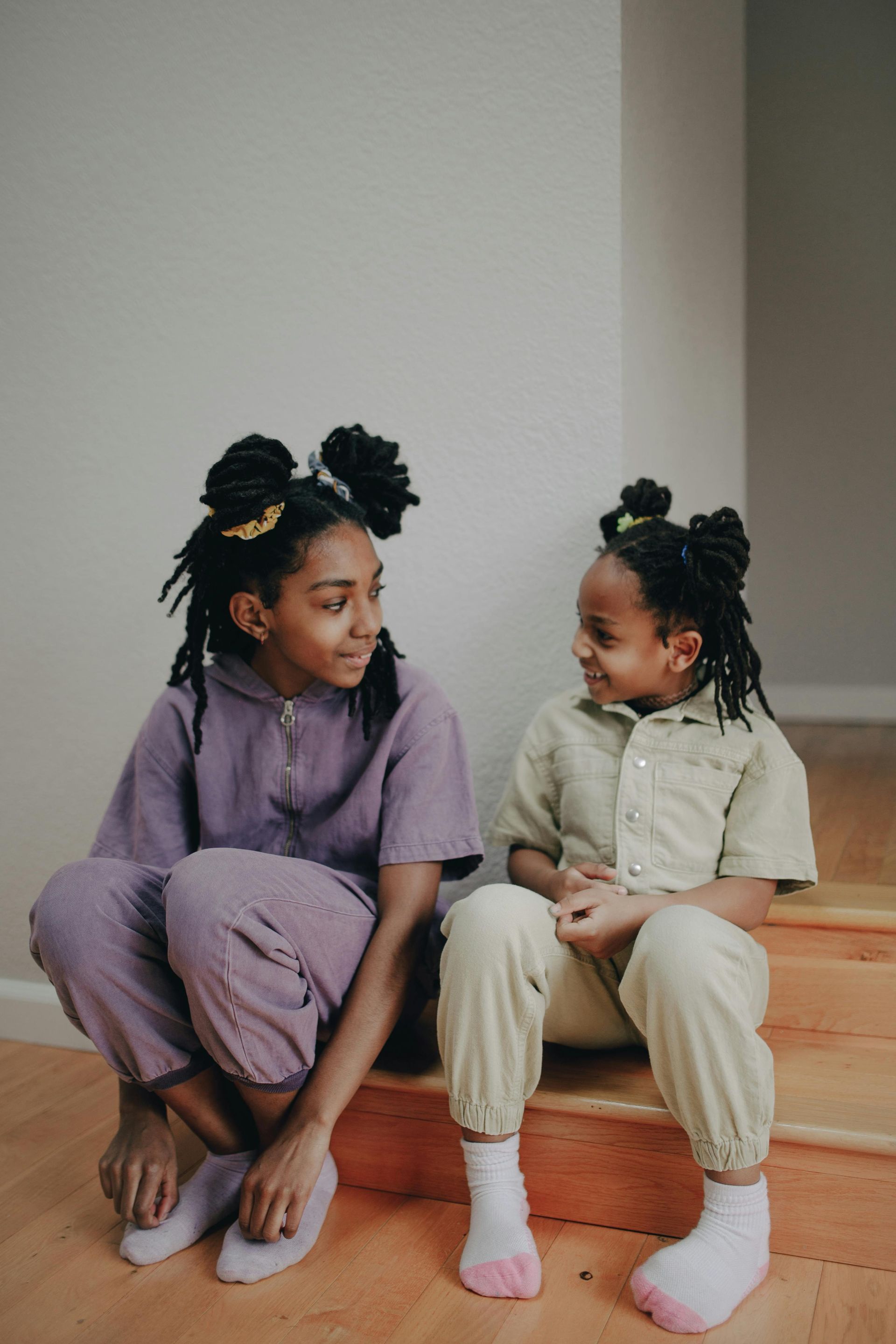 Two Black girls, seated on stairs, look at each other. One wears purple, the other green, both with hair in buns.