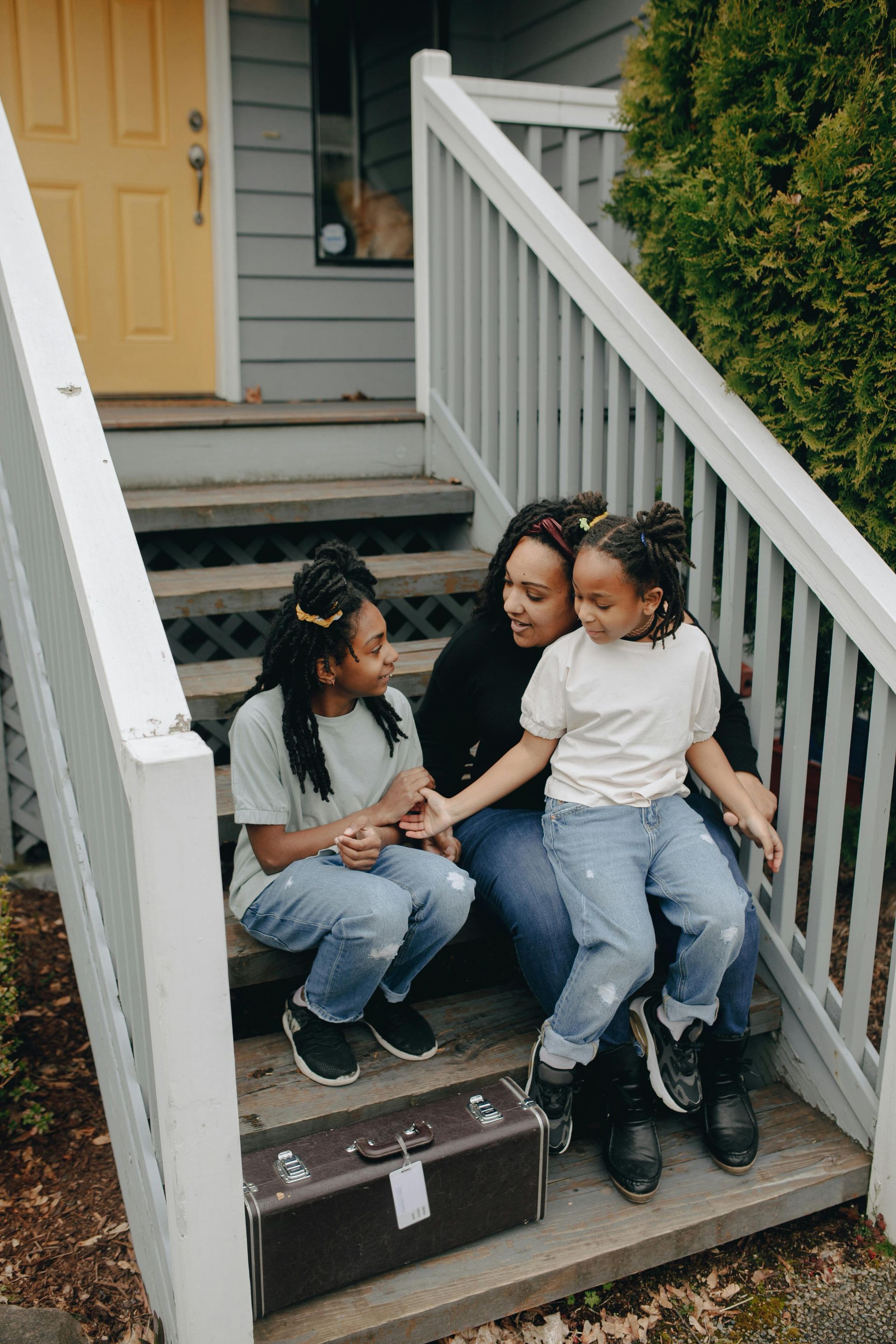 Mother and two children sitting on porch steps, smiling. Vintage suitcase in front of them.