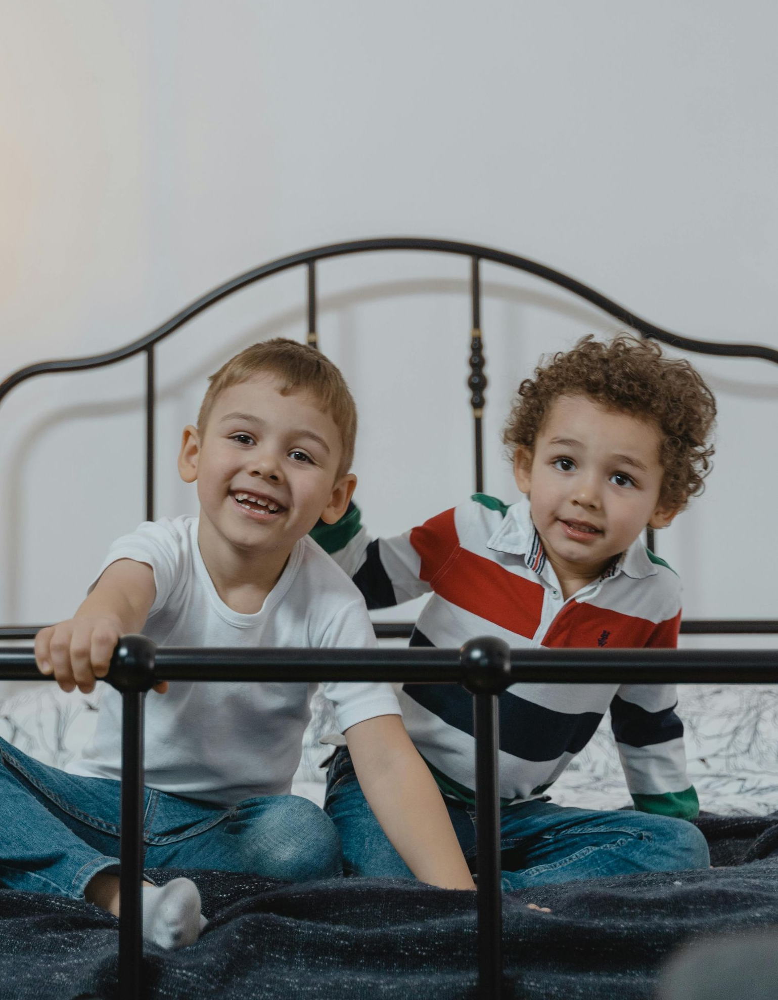Two young boys smiling and sitting on a bed with a black metal headboard.
