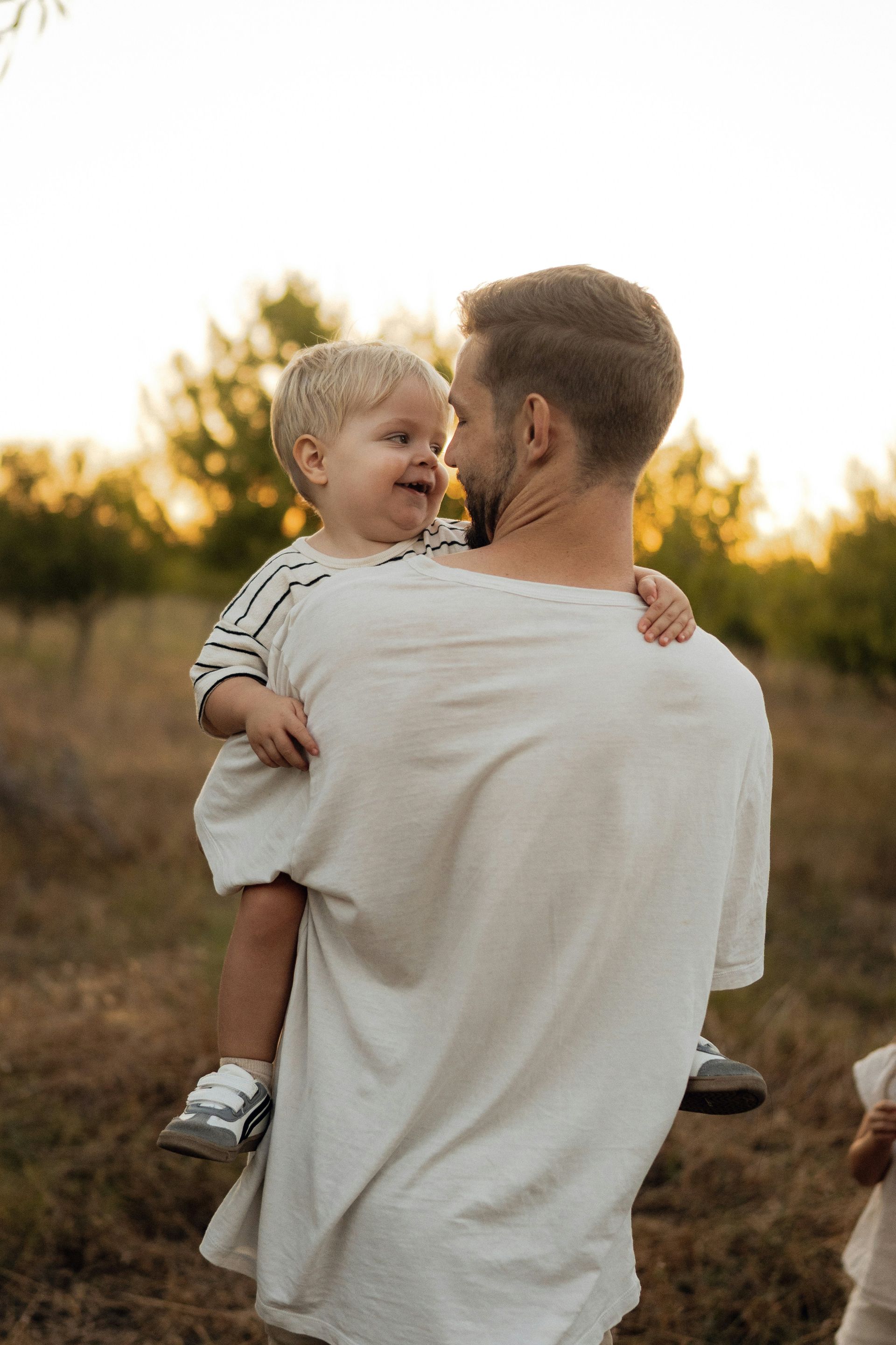 Father holds a laughing toddler in a field. They are smiling at each other; the sunset glows in the background.
