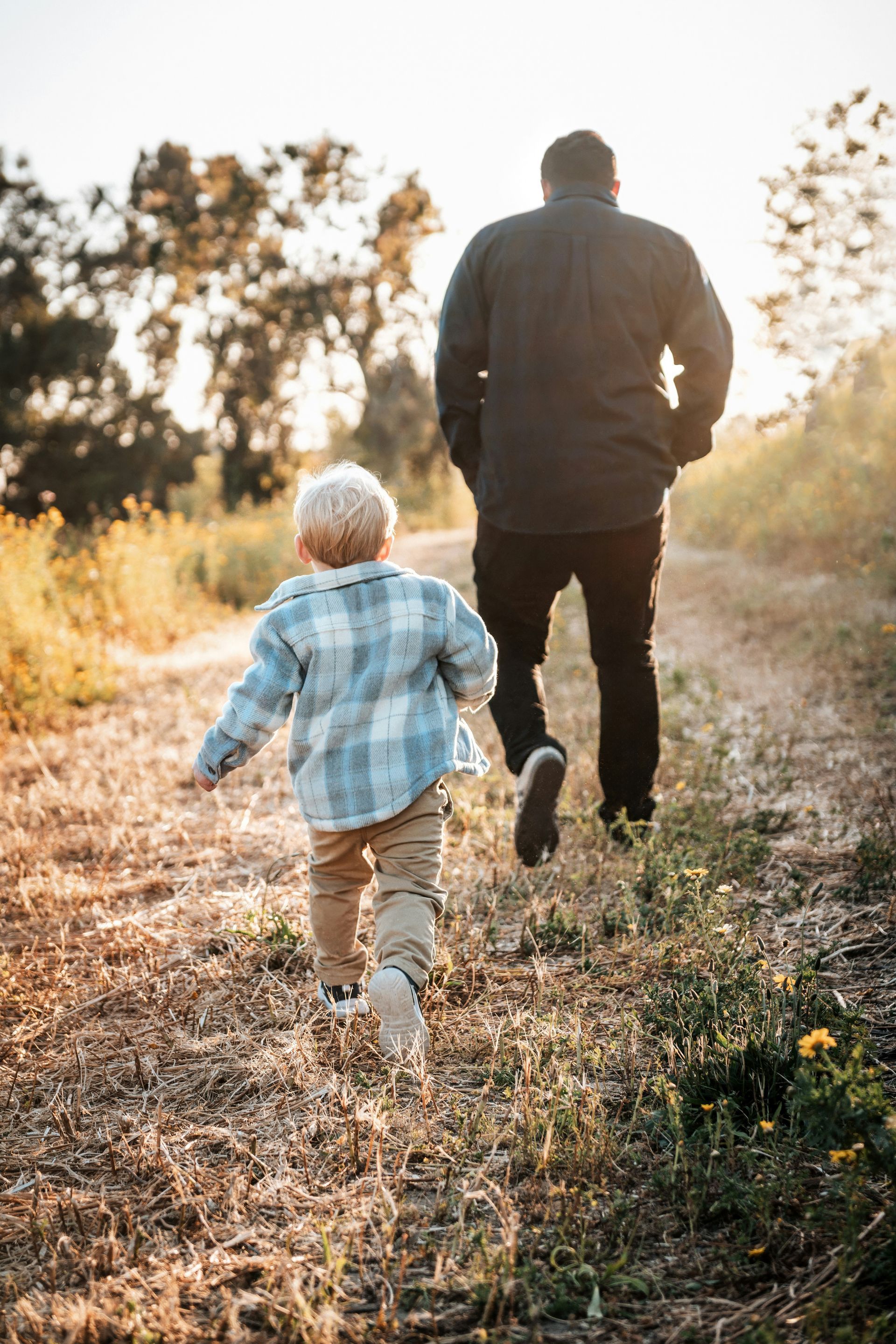 A young boy runs on a dirt path, following a man with his back turned, outdoors at sunset.