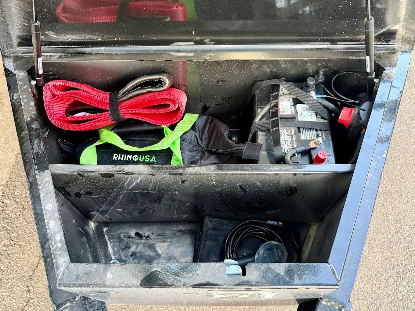Open black trailer toolbox containing recovery gear: tow strap, air compressor, and other tools.