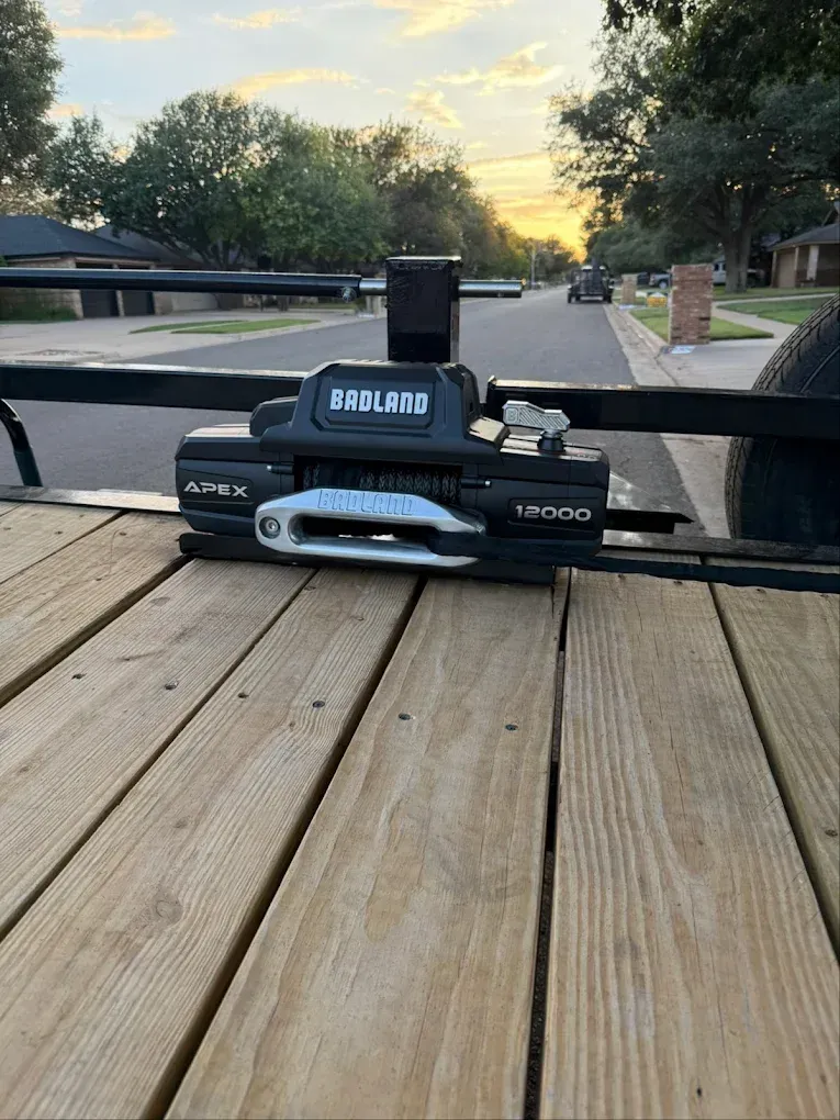 A black Fobland Apex 12000 winch mounted on a wooden trailer bed against a street backdrop.