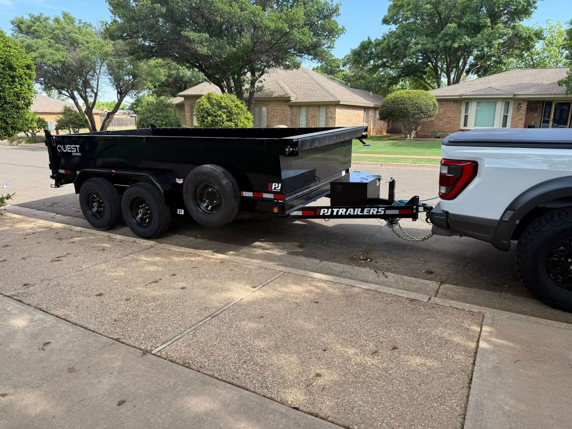 Black dump trailer hitched to a white pickup truck parked on a residential street.