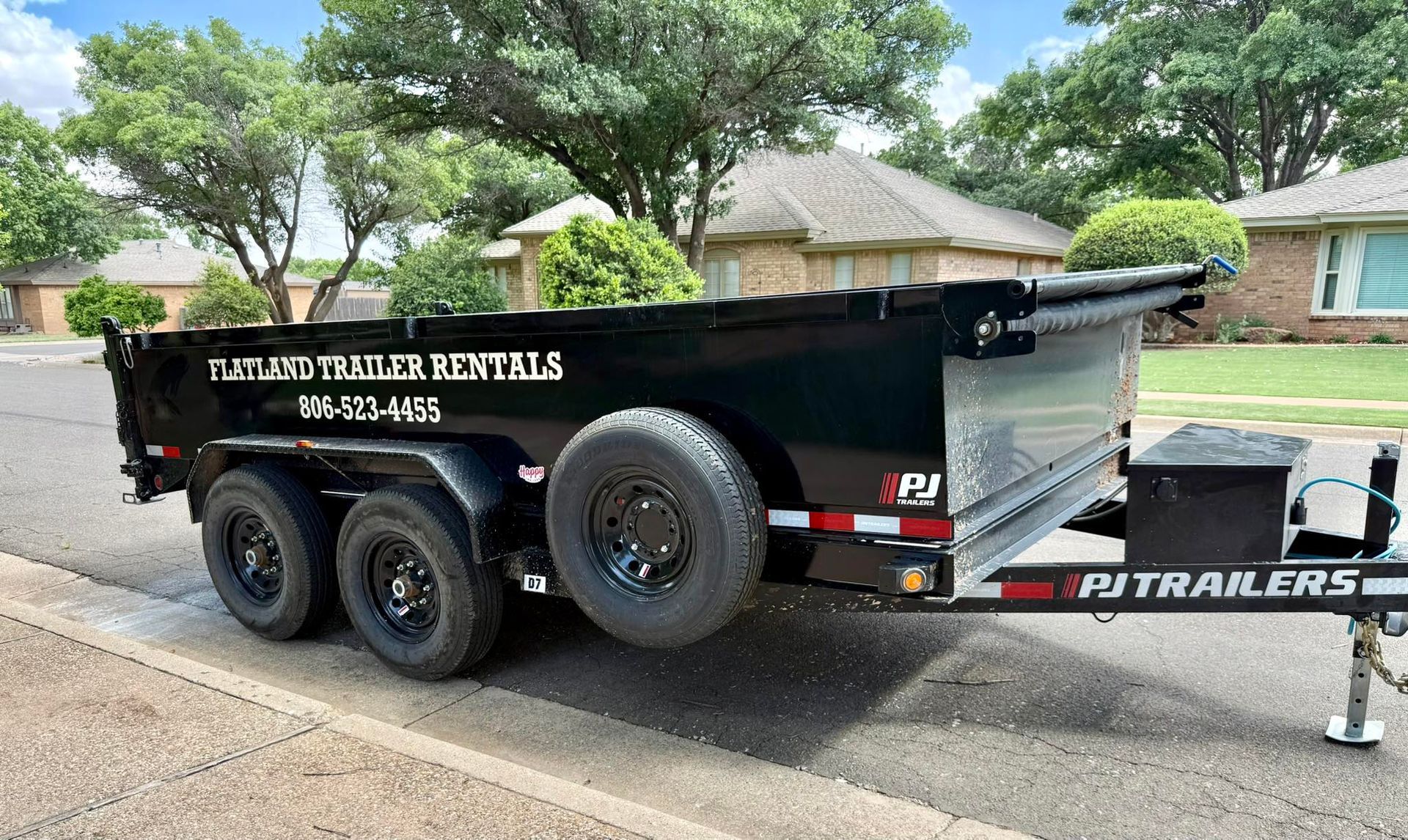 Black PJ Trailers dump trailer parked on a street with a phone number on the side.