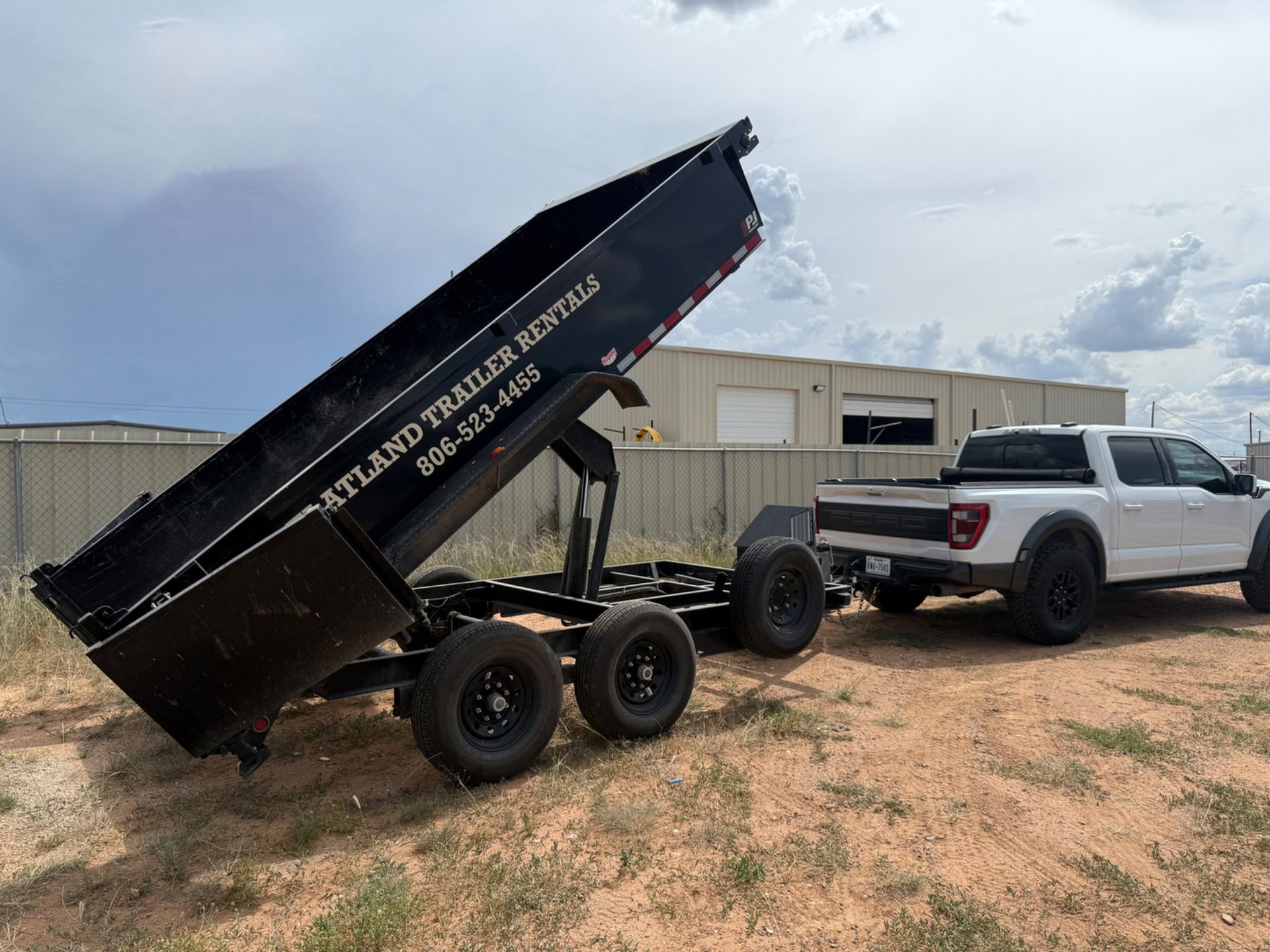 White truck towing a black dump trailer with the bed raised. Cloudy sky in the background.