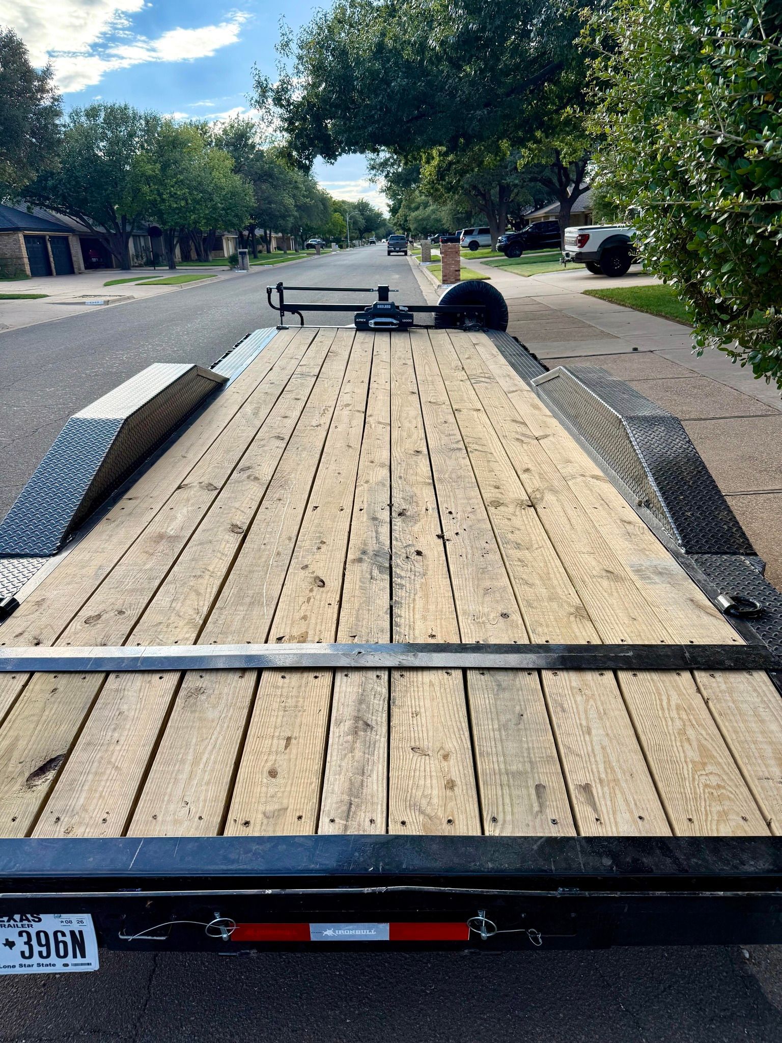 Flatbed trailer parked on a residential street. Wooden deck, black frame, and a black hitch.