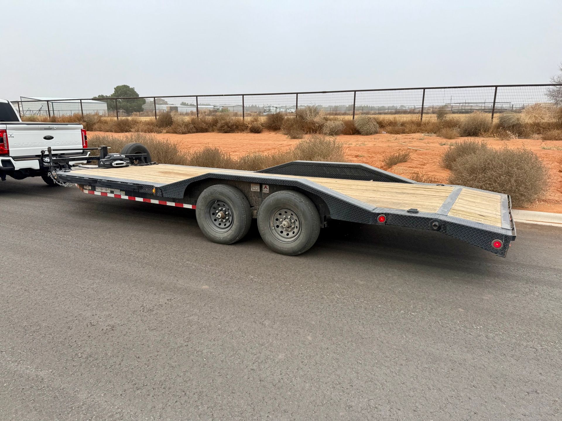 Truck towing a black flatbed trailer on asphalt road with beige landscape and a fence in the background.