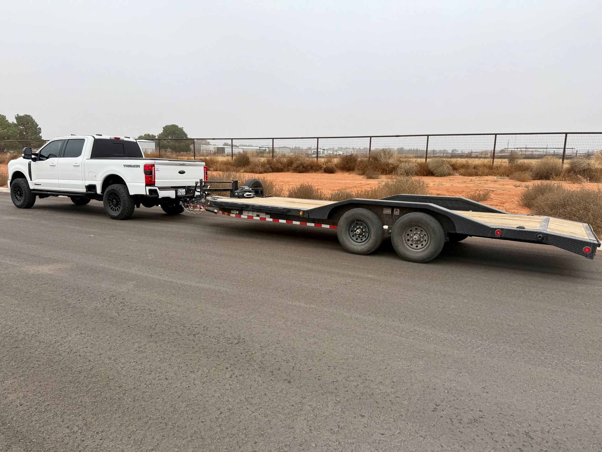 White pickup truck towing a black trailer on asphalt road with a fence and sparse vegetation in the background.