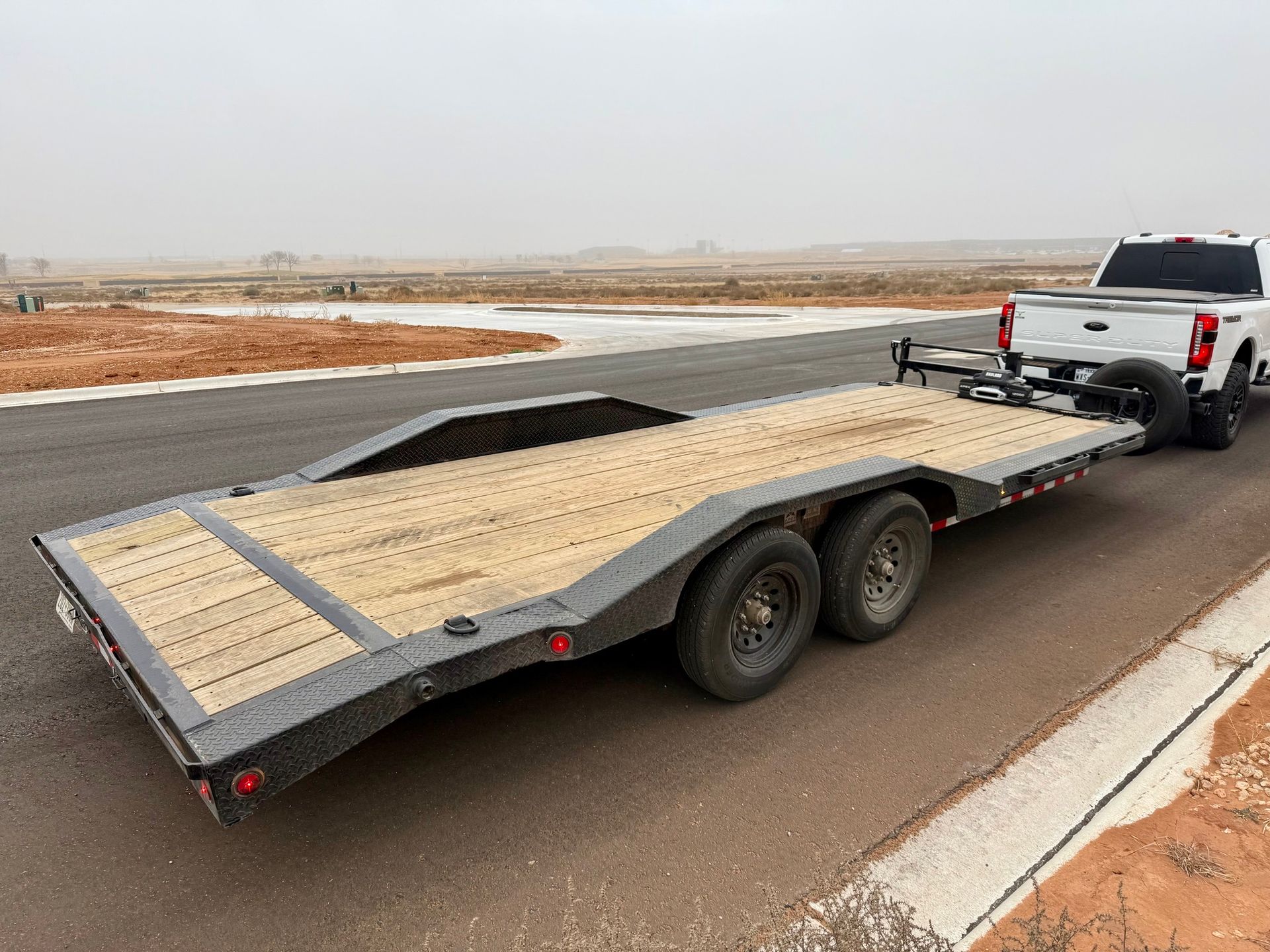 White pickup truck towing a black flatbed trailer on an asphalt road in a desert landscape.