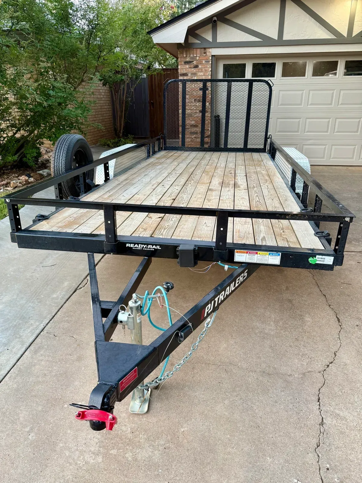 Black utility trailer on a driveway. Light wood planks, black frame. Spare tire, hitch, and ramp.