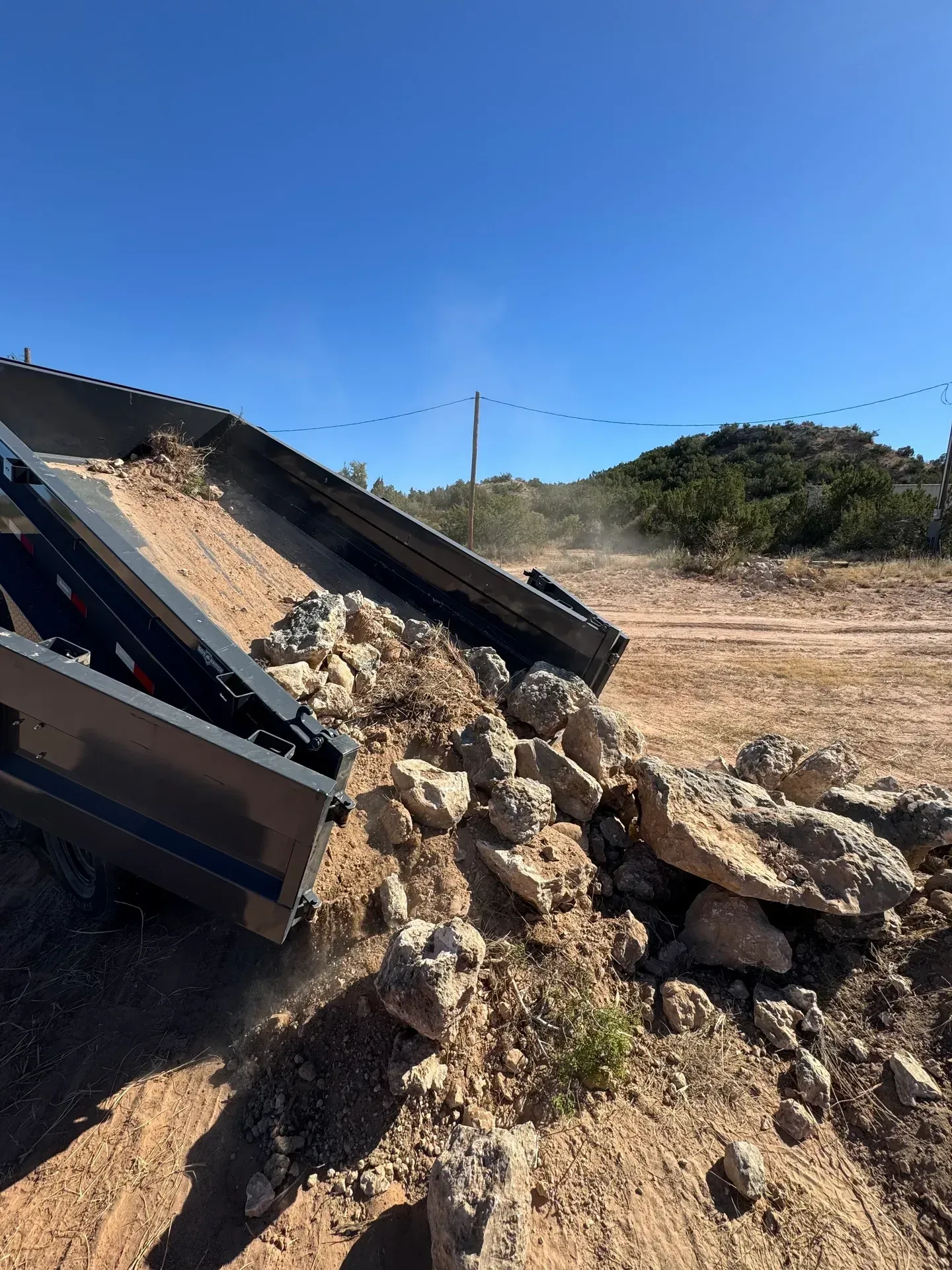 Dump truck unloading dirt and rocks onto a dusty rural field under a bright blue sky.