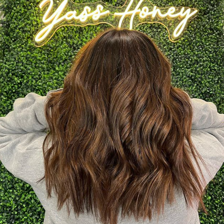 A woman with long brown hair is standing in front of a neon sign that says yass honey.