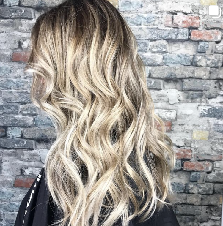 A woman with long blonde hair is standing in front of a brick wall.