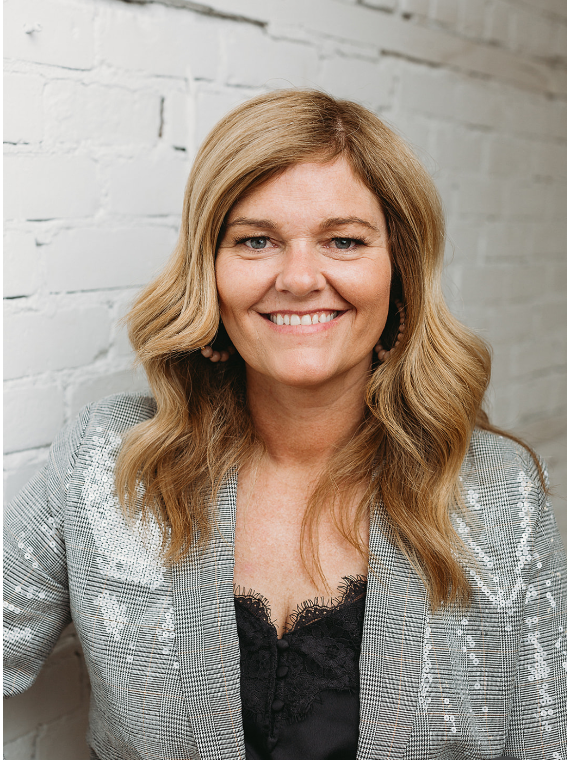 A woman is smiling and leaning against a white brick wall.