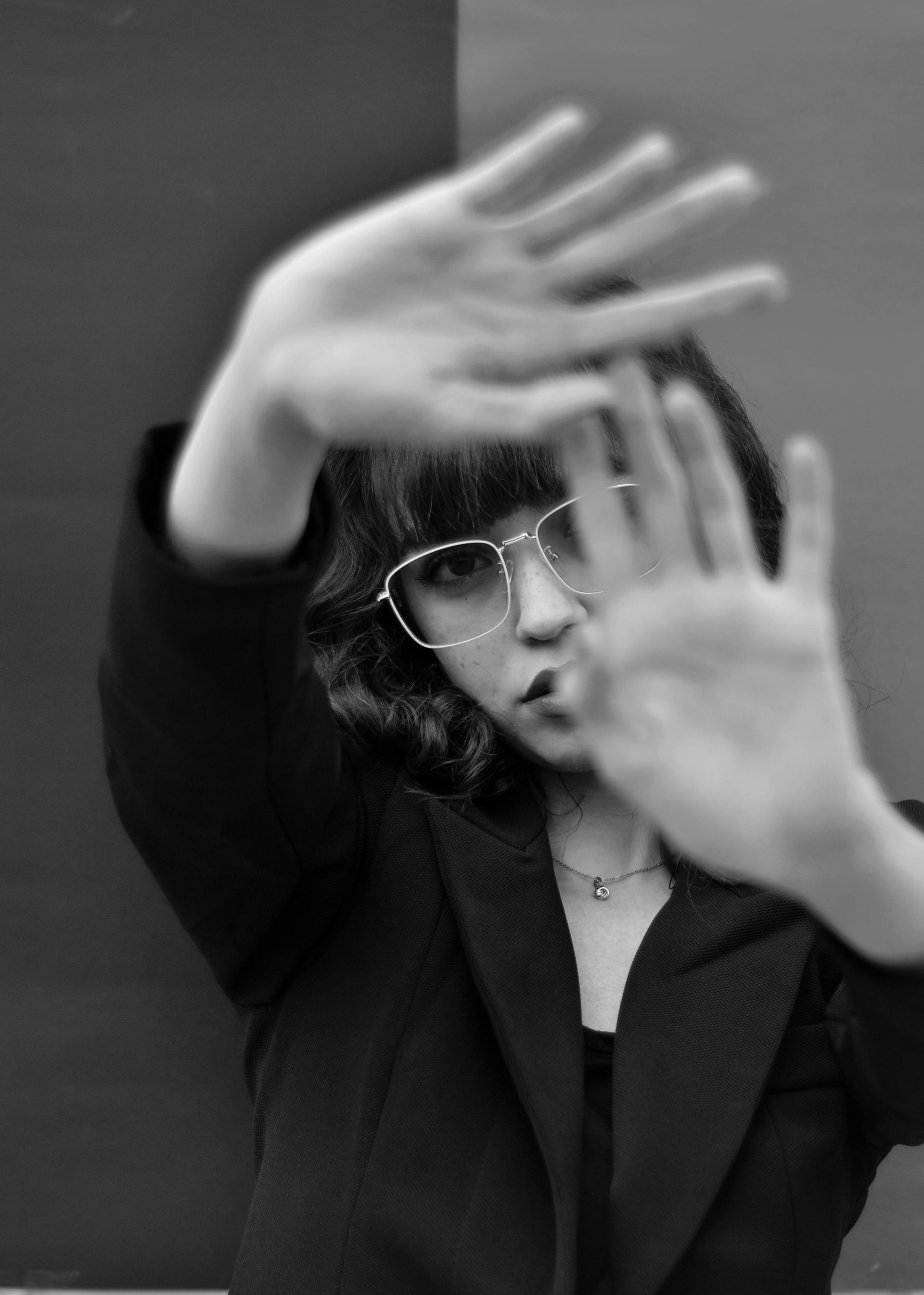 Person in glasses holds hands up toward the camera, blurry face, against a two-tone background.