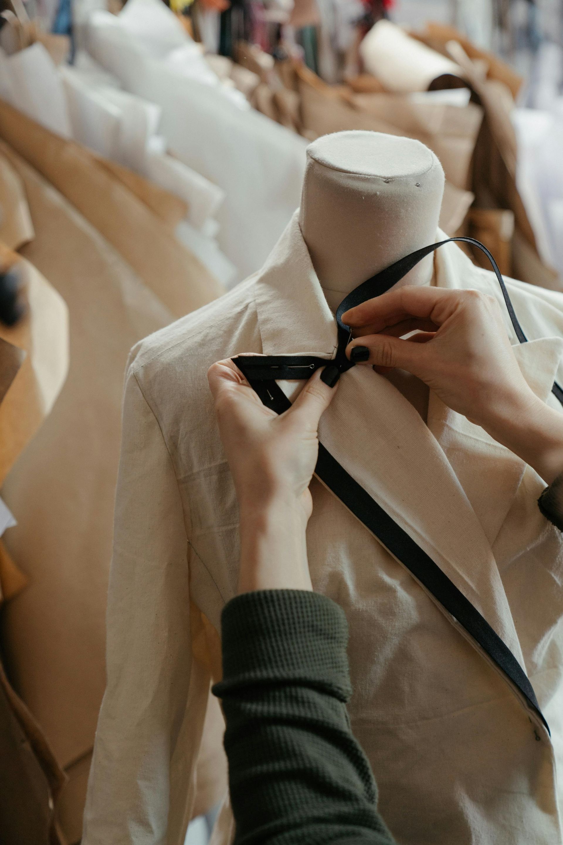 Hands adjusting a black ribbon on a cream-colored blazer on a mannequin in a studio.