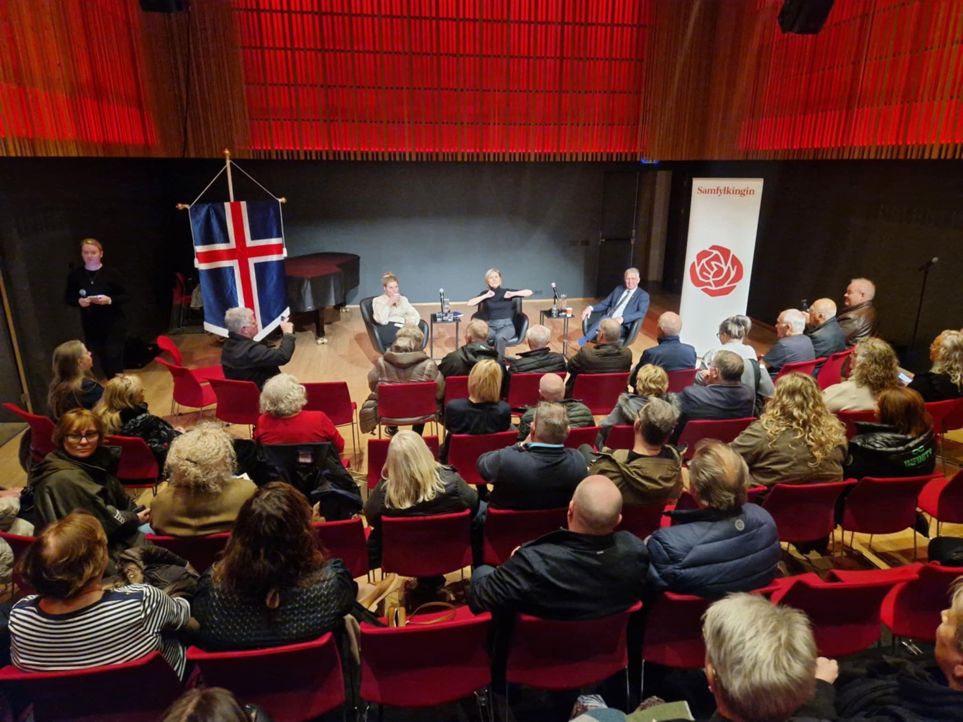 Audience listens to a panel discussion in Berg hall, with an Icelandic flag and Samfylkingin banner on stage.