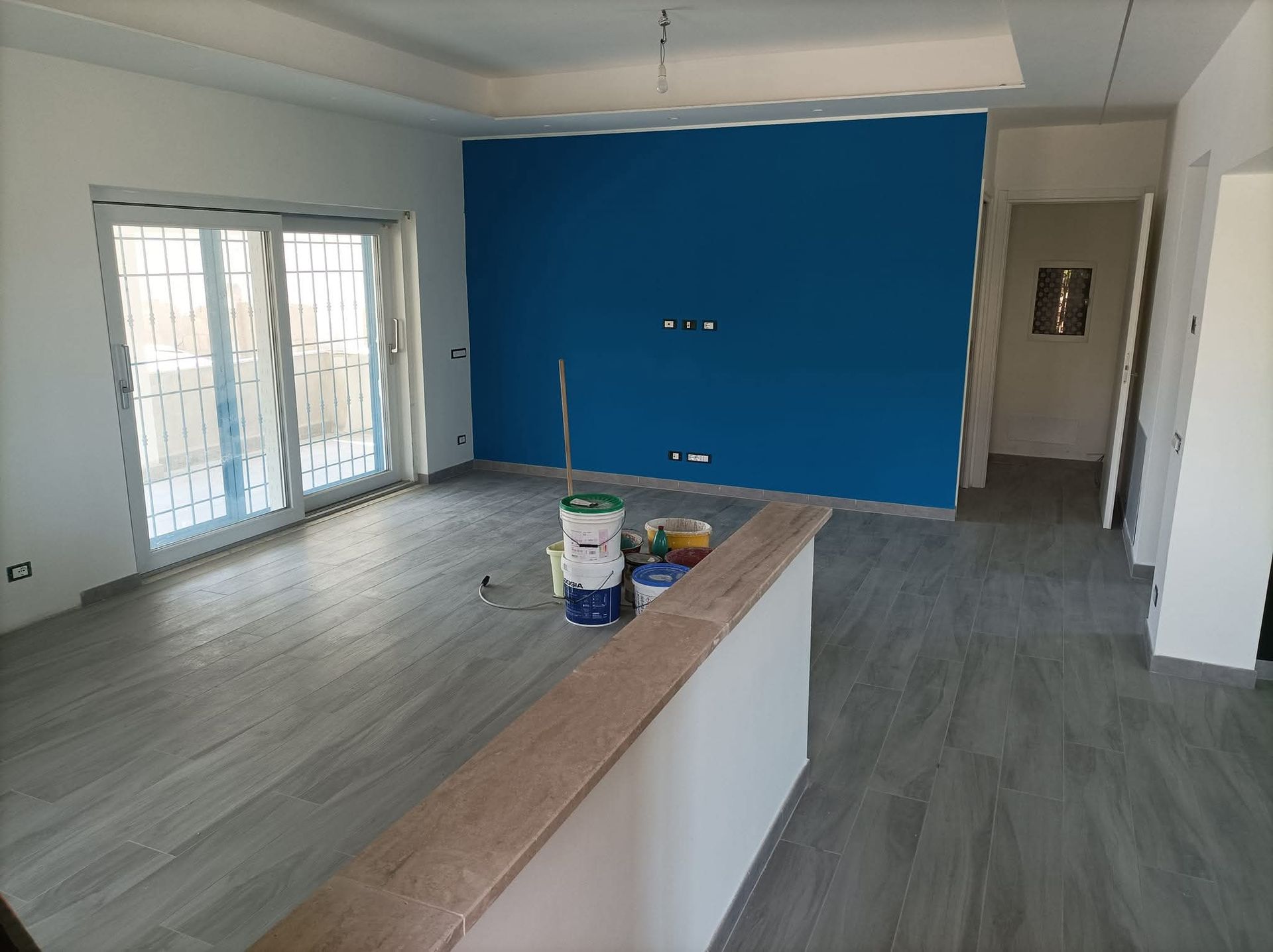 Stanza in fase di ristrutturazione con una parete blu, pavimento grigio e porte scorrevoli aperte. Barattoli di vernice sul pavimento.