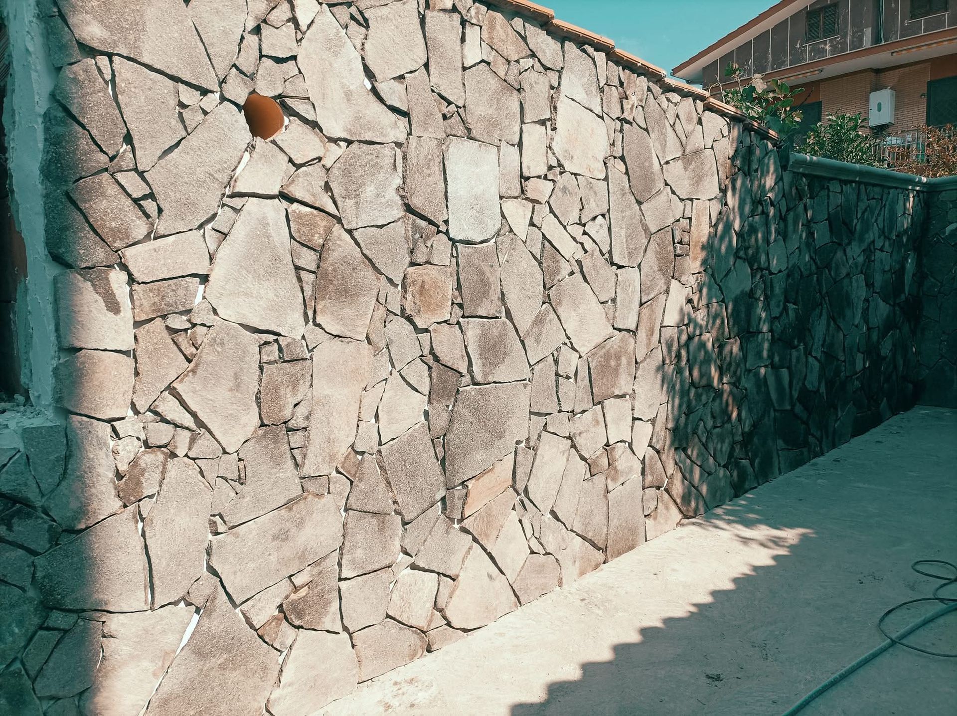 Muro di pietra costituito da pietre grigie irregolari. Sono visibili una lastra di cemento e un edificio.