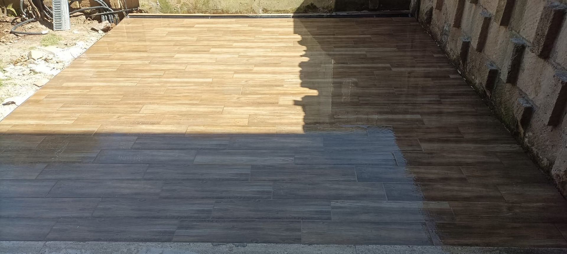 Terrazza in legno, in parte bagnata, in parte asciutta. La parte asciutta è marrone chiaro, quella bagnata è grigio scuro. Muro di mattoni a destra.