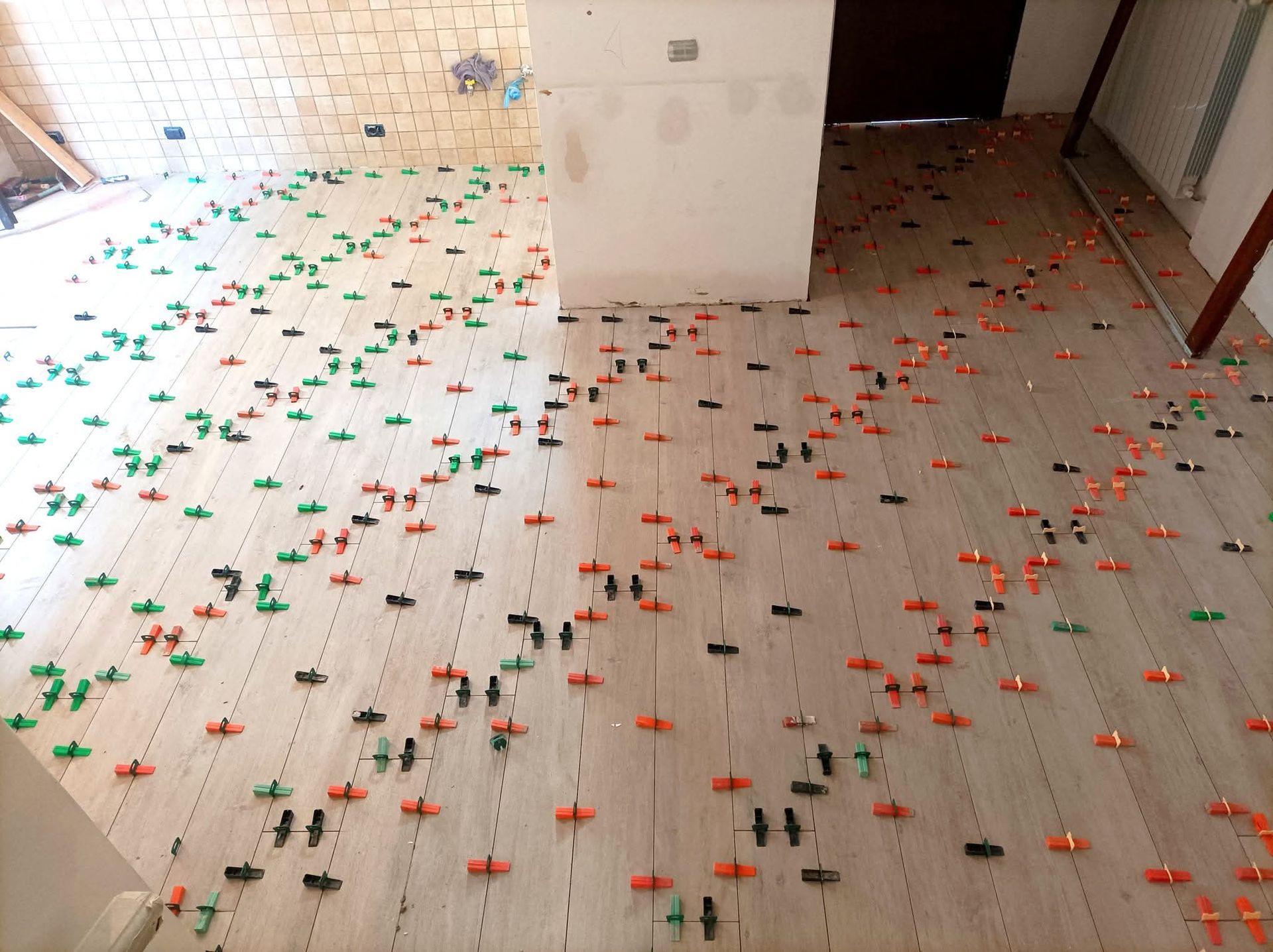 Piastrelle in fase di installazione, con graffe di livellamento in uso. Graffe verdi, arancioni e nere punteggiano il pavimento chiaro.