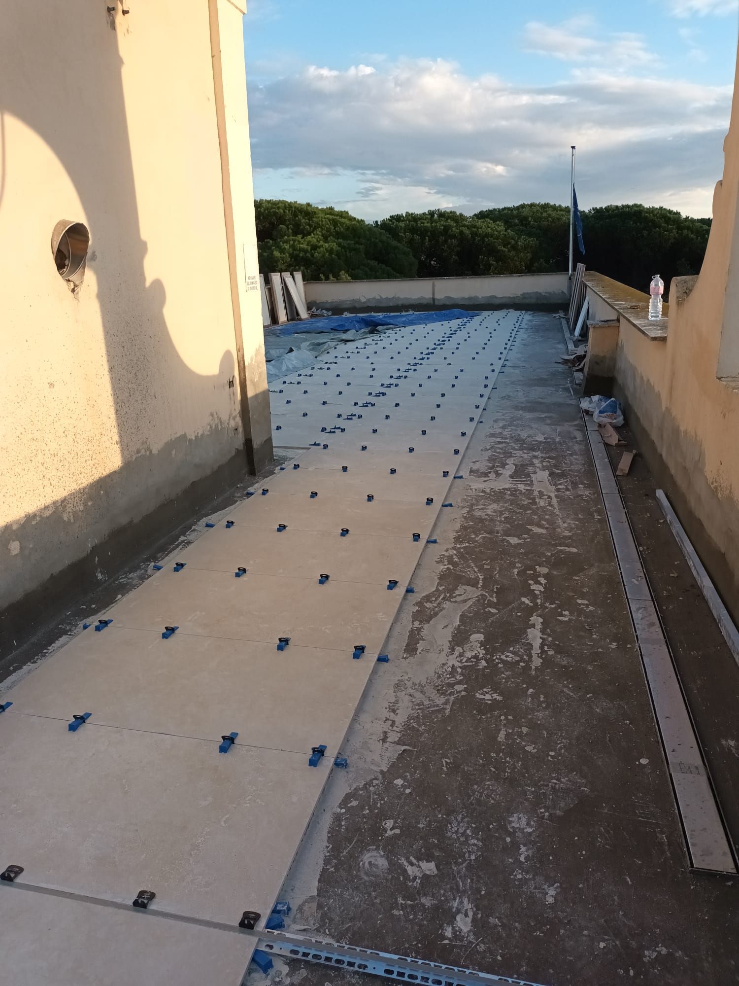 Tetto piastrellato in costruzione con distanziatori di livellamento in posizione, tegole beige, cielo azzurro e alberi sullo sfondo.