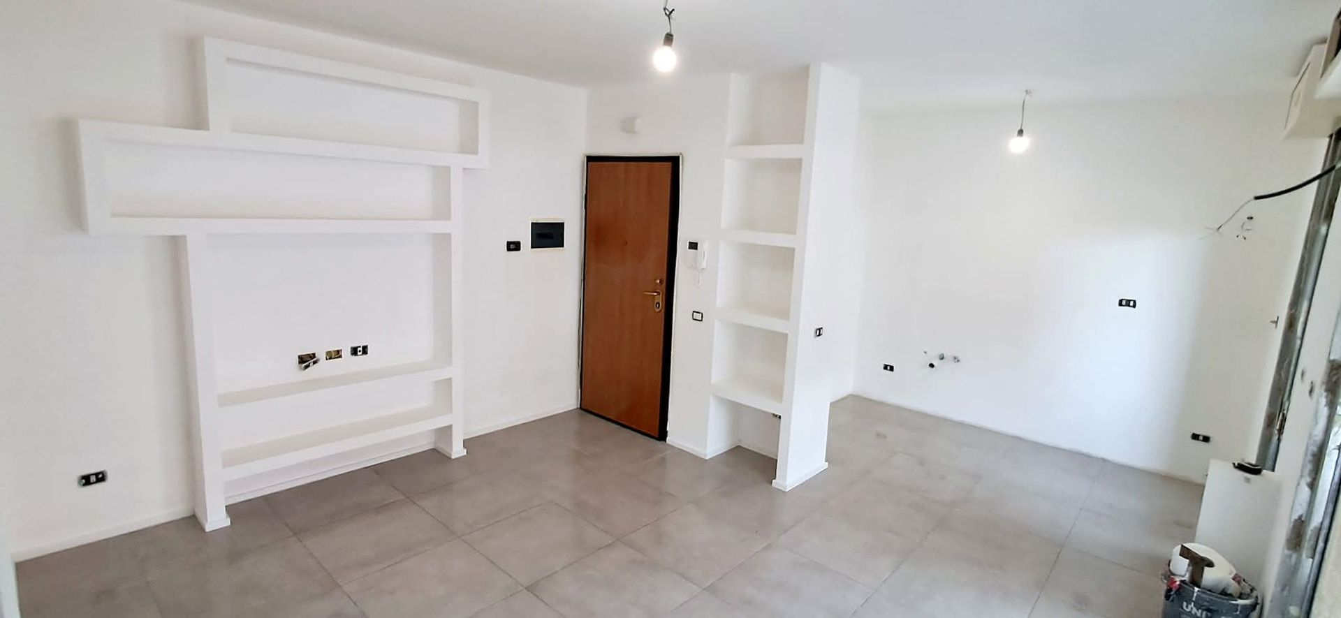 Stanza vuota con scaffali a muro e una porta marrone. Pavimento piastrellato grigio e pareti bianche.