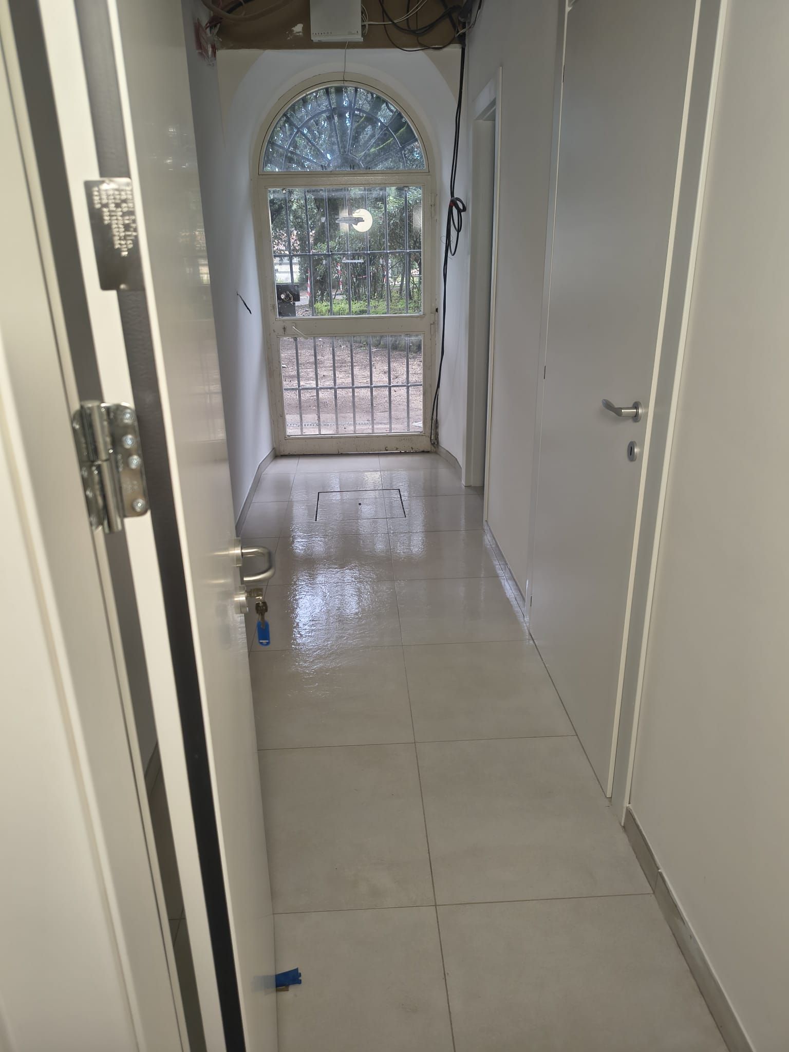 Corridoio stretto con porte bianche e pavimento piastrellato chiaro; una finestra ad arco in fondo si apre sul verde.