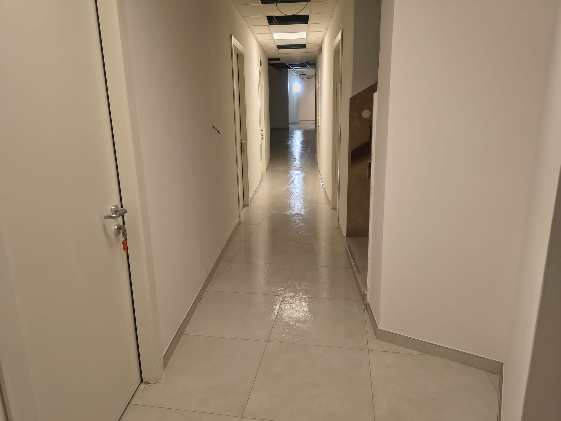 Corridoio lungo, stretto e bianco con porte chiuse su entrambi i lati, pavimento piastrellato lucido, illuminazione dall'alto.