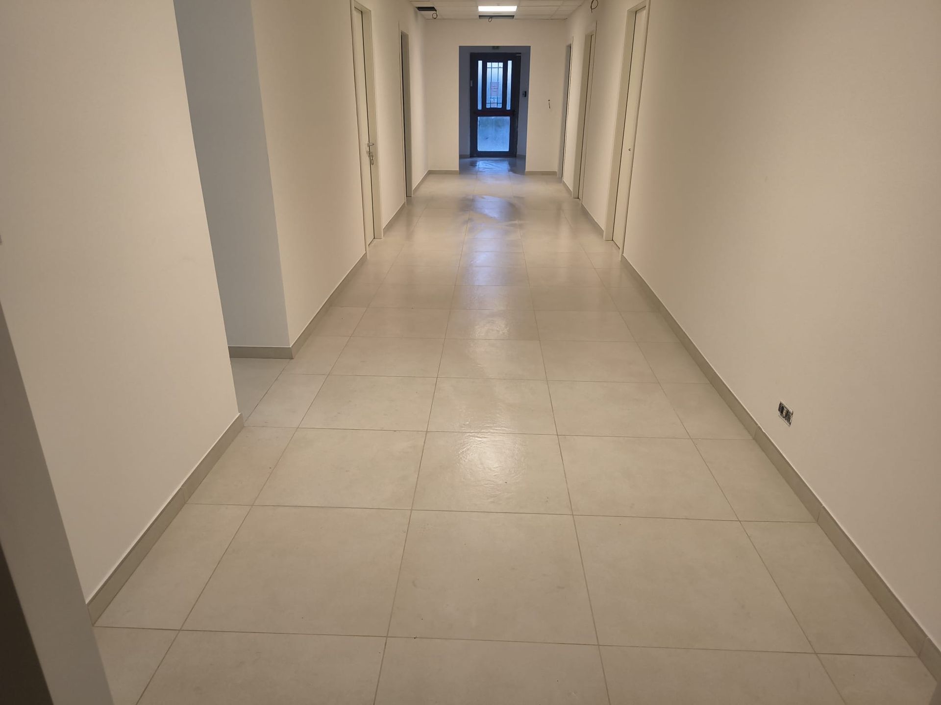 Un lungo corridoio vuoto, con pareti bianche e pavimento piastrellato. Diverse porte chiuse fiancheggiano le pareti.