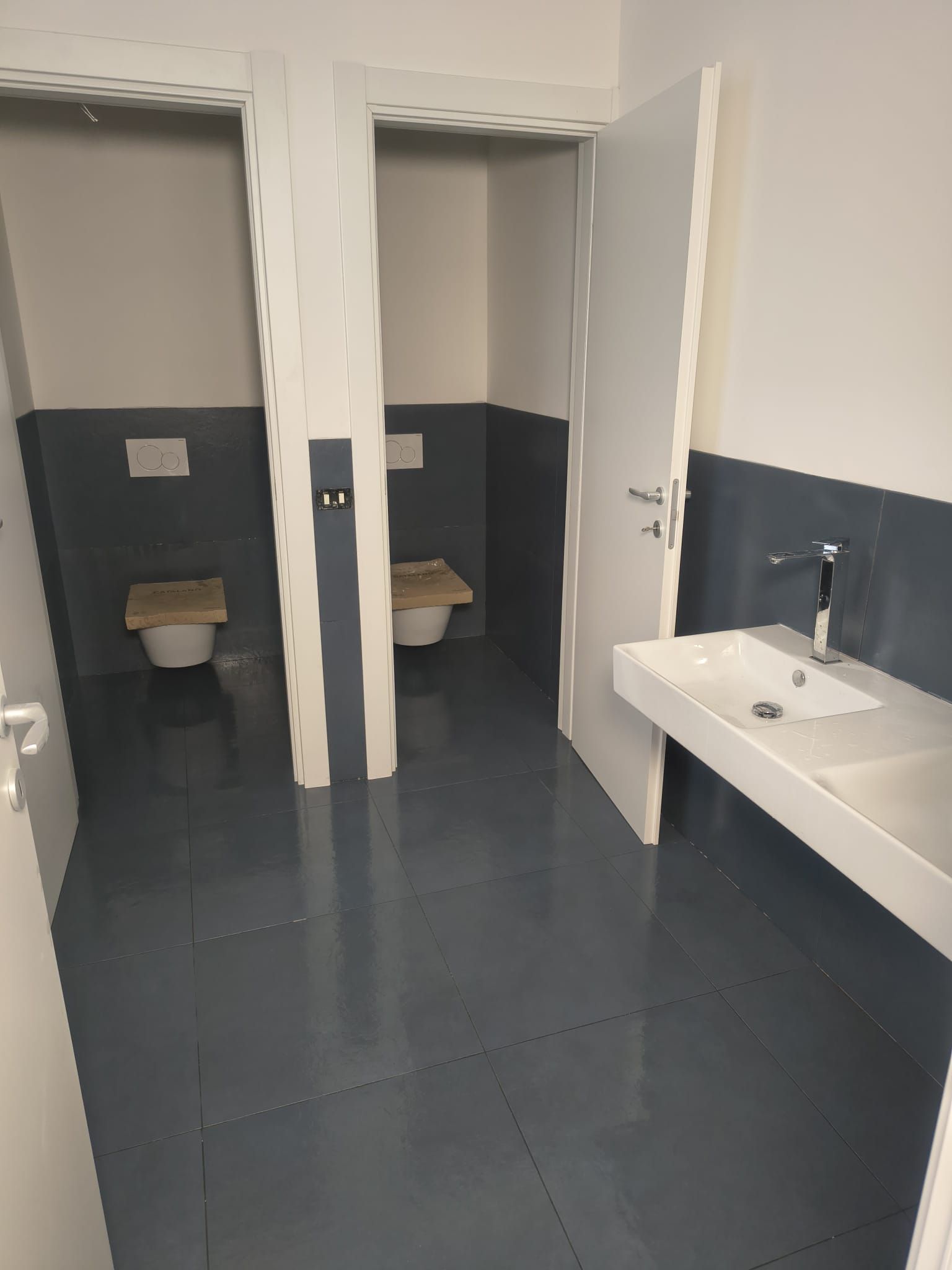 Bagno pubblico con due cabine, un lavandino e pareti e pavimento grigi.