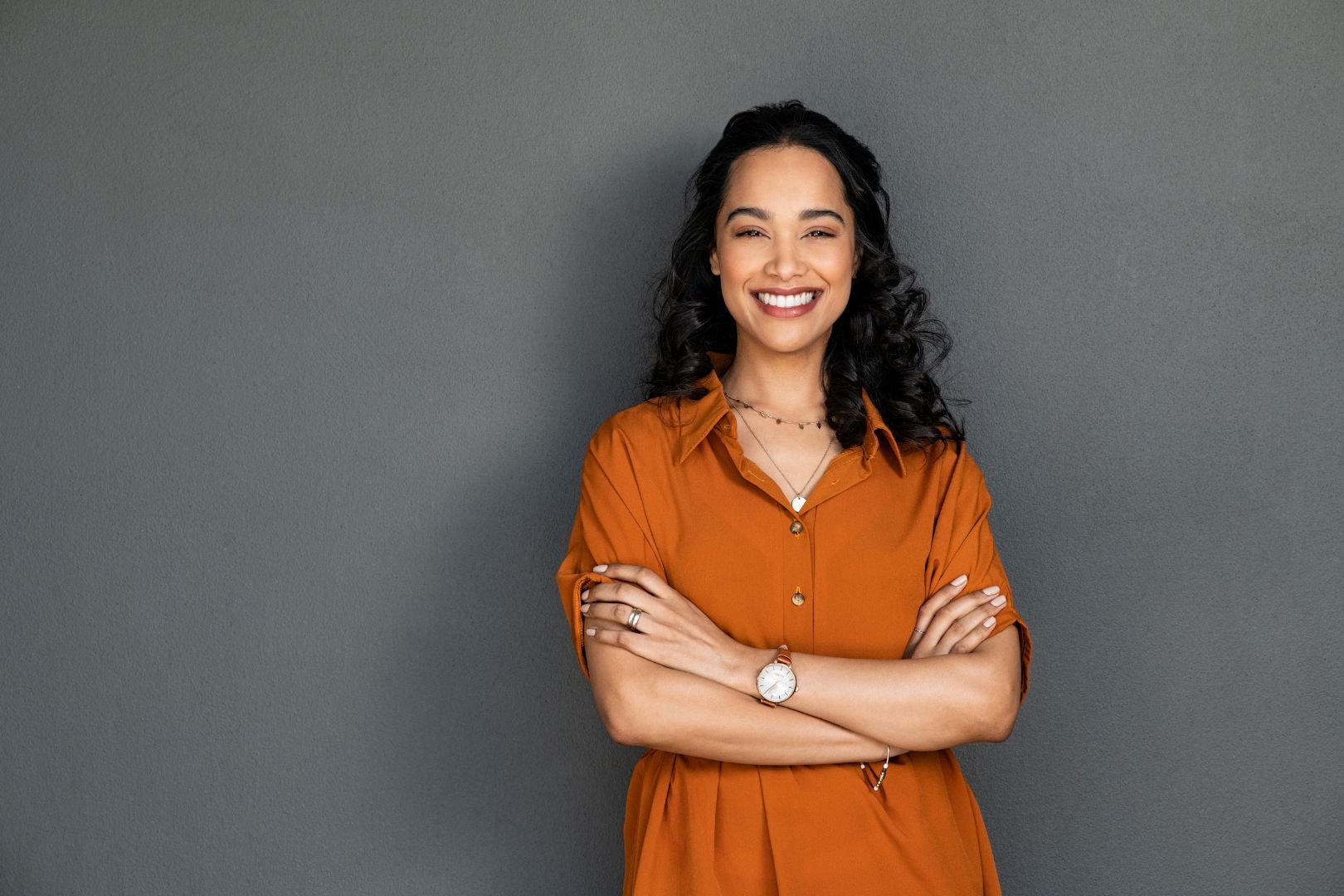 Woman with crossed arms smiles against a gray wall, wearing orange shirt and wristwatch.