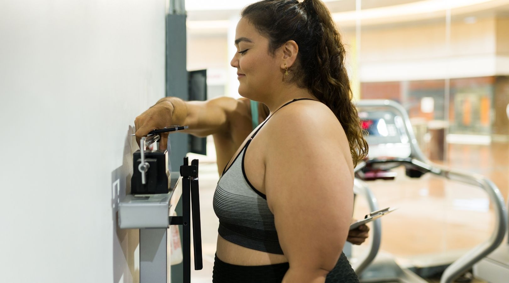 Woman adjusting weight on a scale at a gym, wearing workout clothes, smiling.