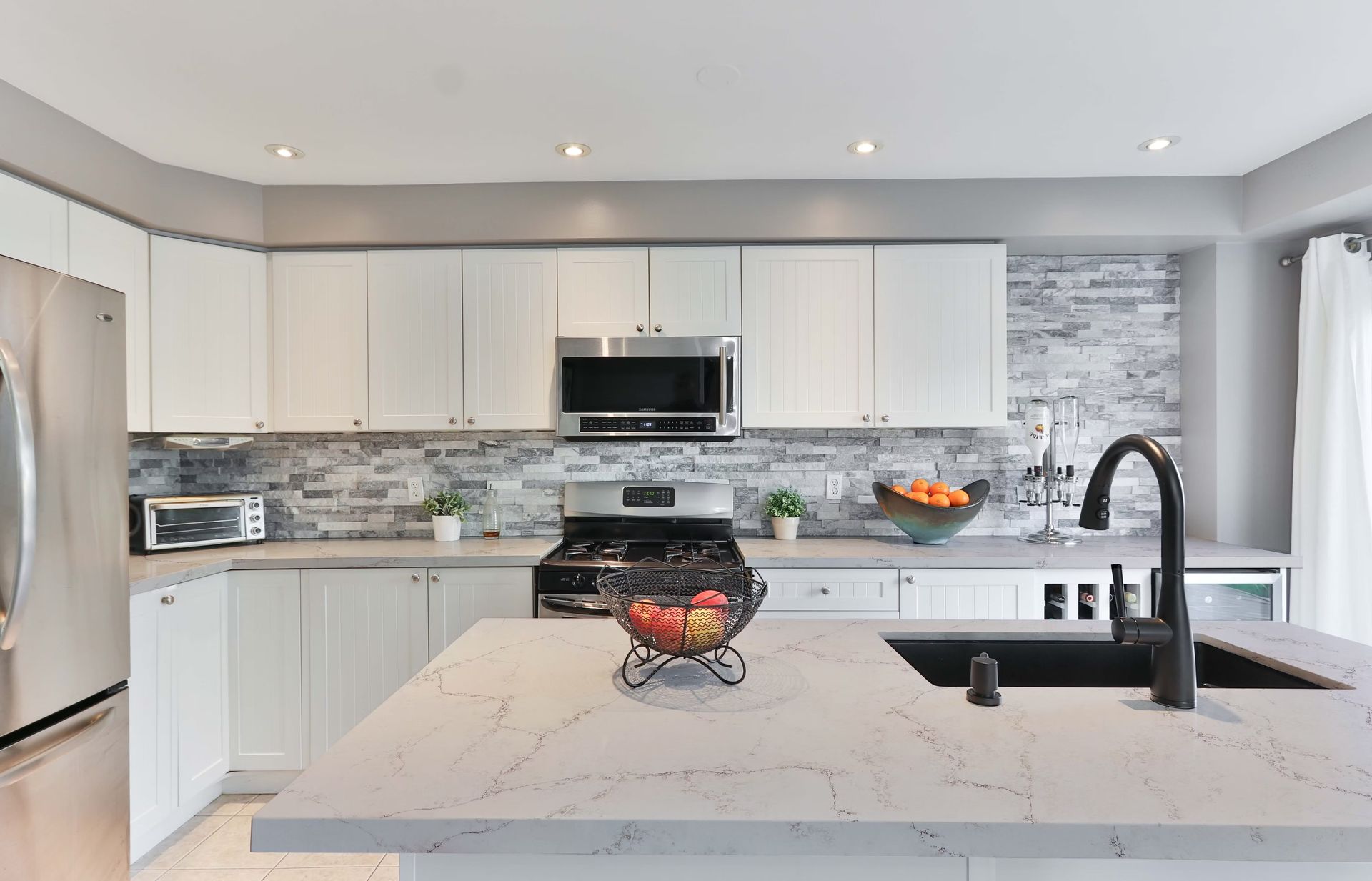 Modern white kitchen with stainless steel appliances, stone backsplash, and island with black faucet.