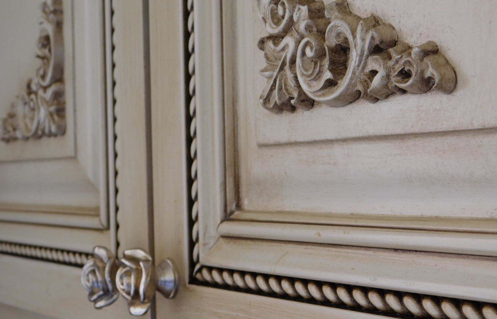 Cream-colored cabinet doors with ornate carvings and rope detailing, silver knobs.