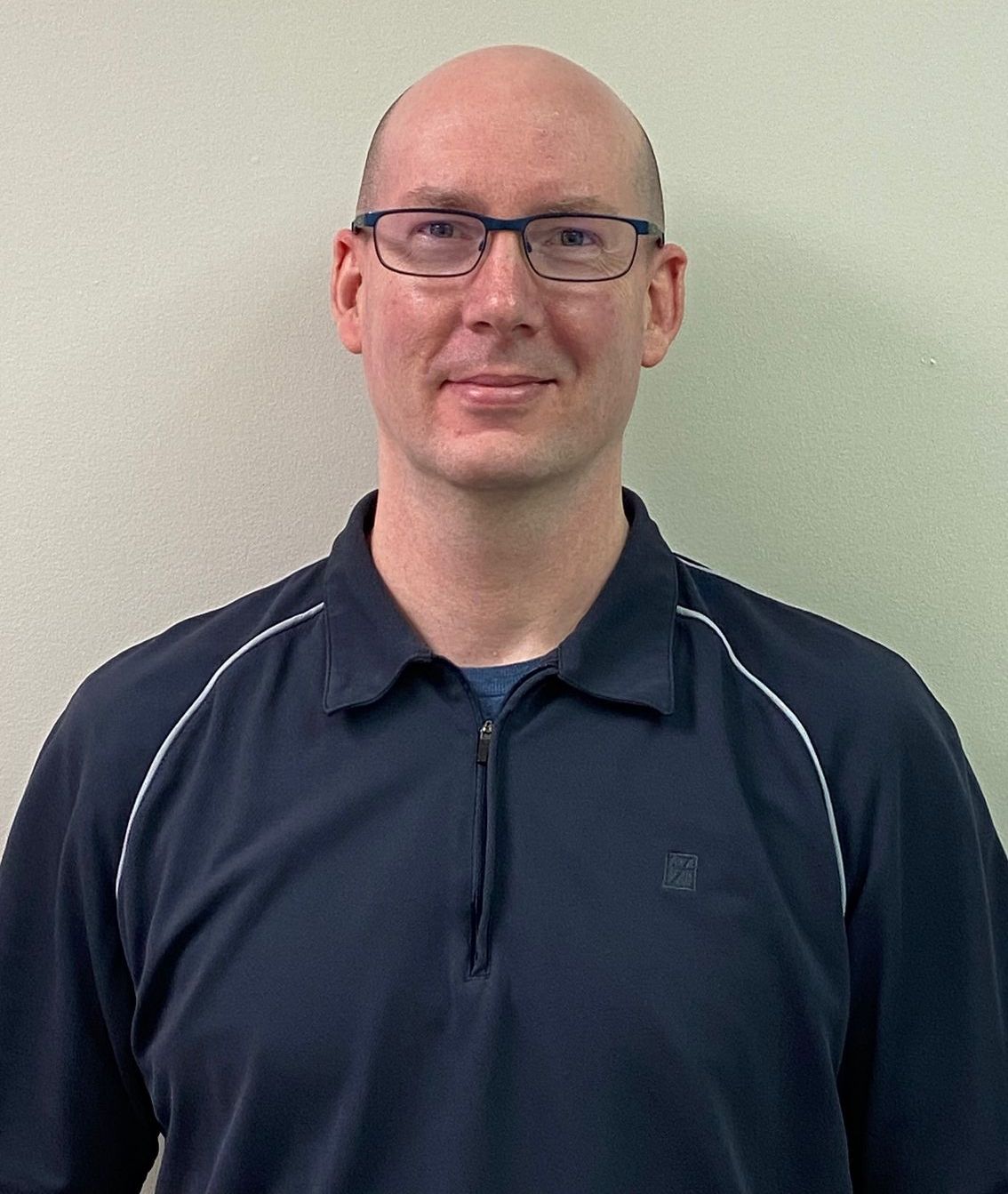 A bald man wearing glasses and a blue shirt is standing in front of a white wall.