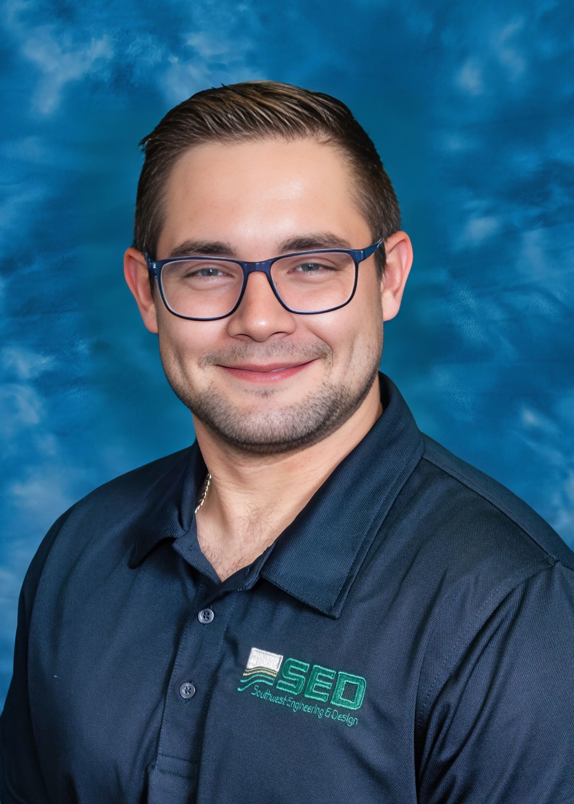 Man with glasses and a smile, wearing a dark blue polo shirt, in front of a blue backdrop.