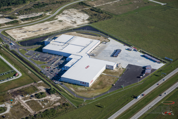 Aerial view of a large white building with loading docks and a parking lot, surrounded by green fields and roads.