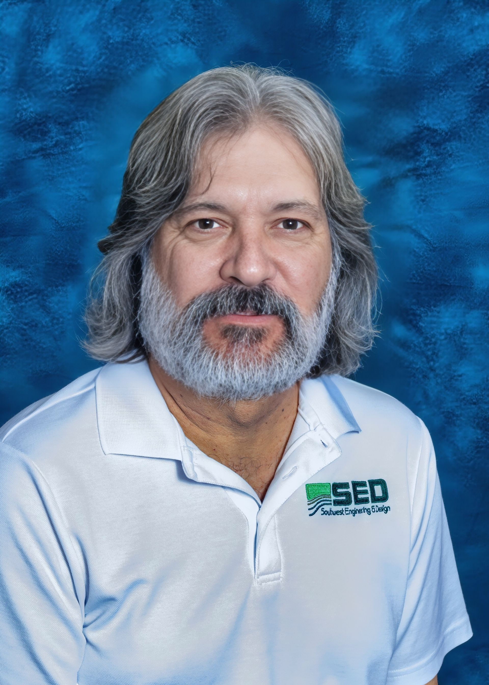 Man with gray hair and beard wearing a white polo shirt with a logo, against a blue background.