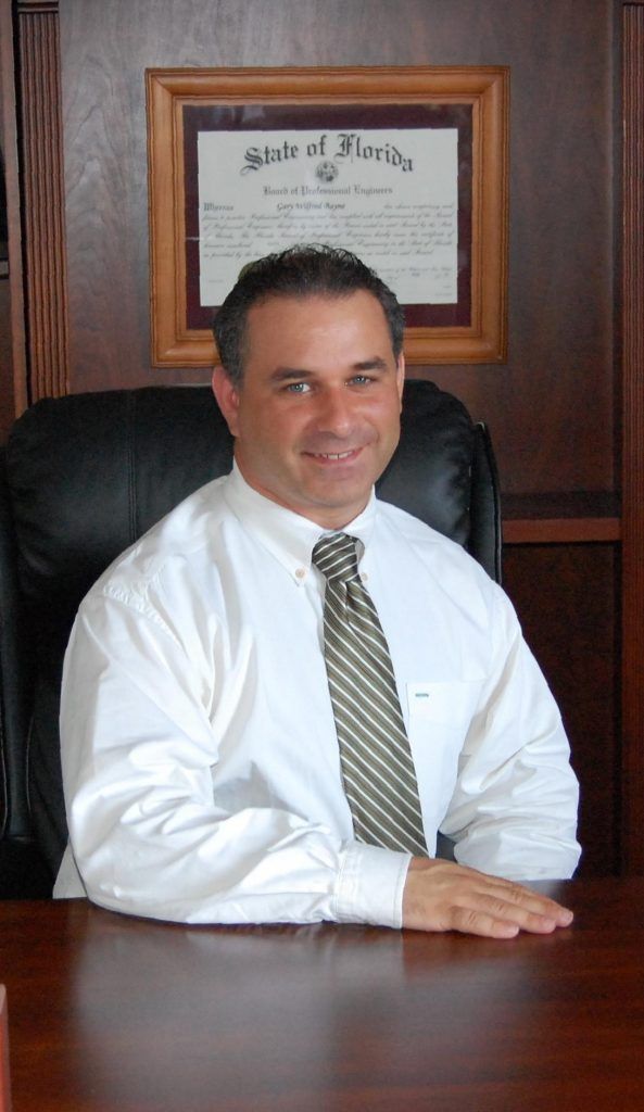 Man in white shirt and tie sitting at desk, smiling. Behind him is a framed certificate.
