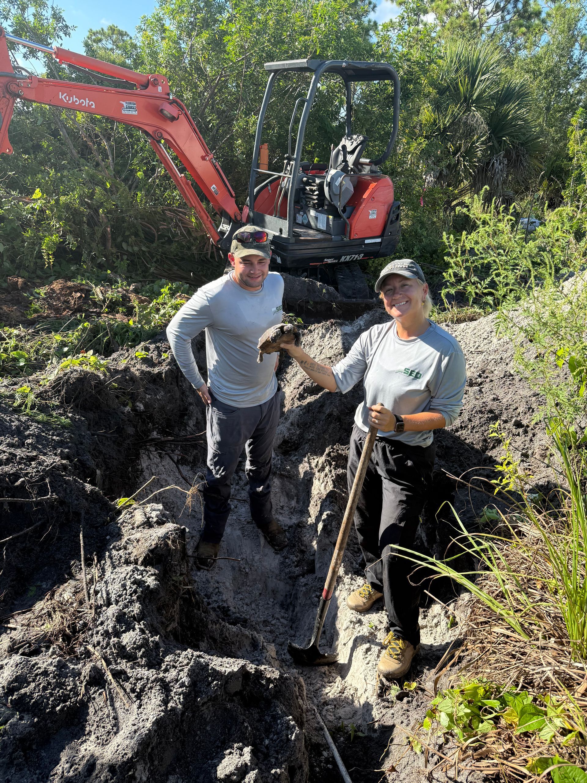 Two people by a small excavator, digging a trench outdoors.