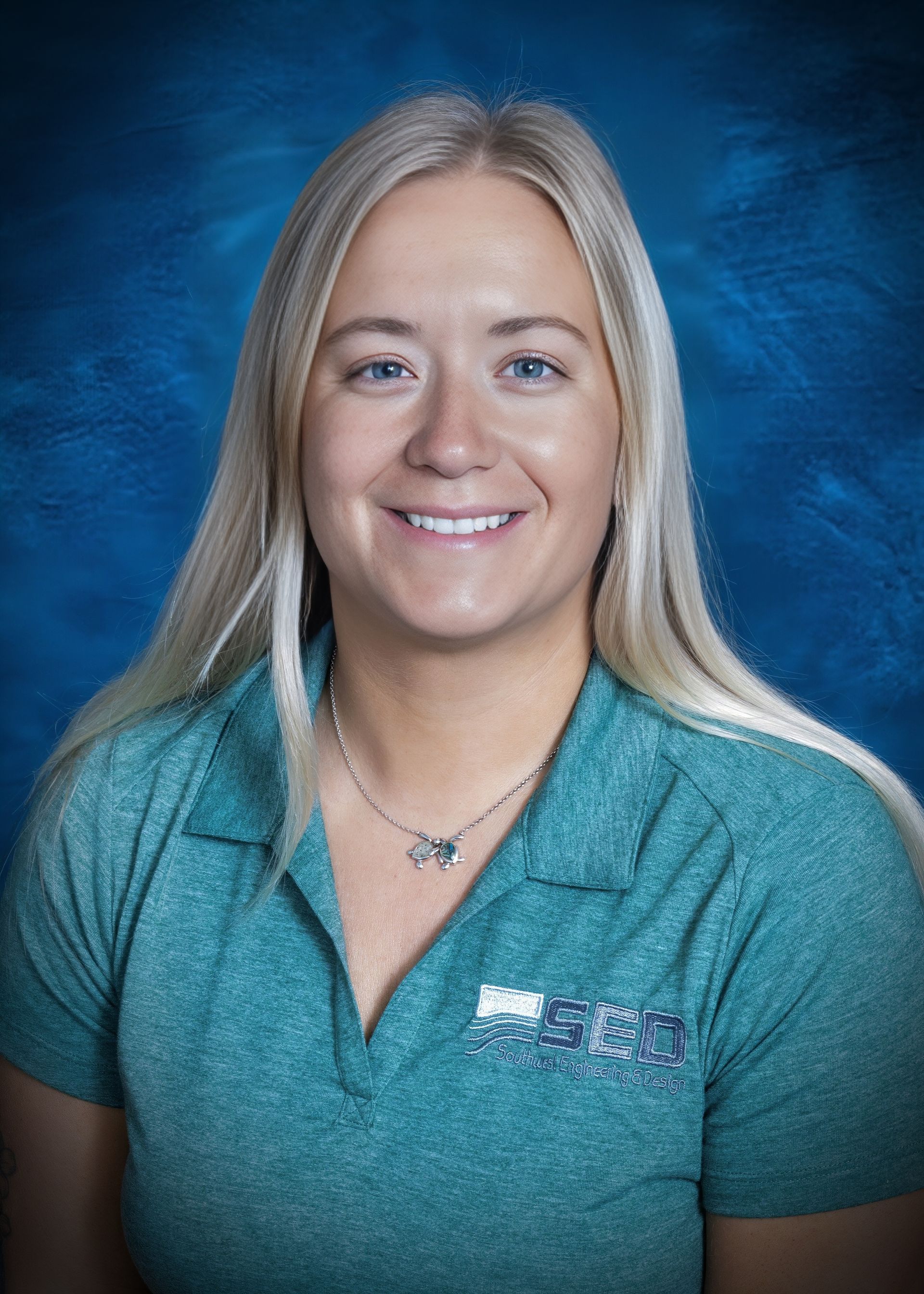 Blonde woman with a smile wearing a teal polo shirt, necklace, against a blue backdrop.