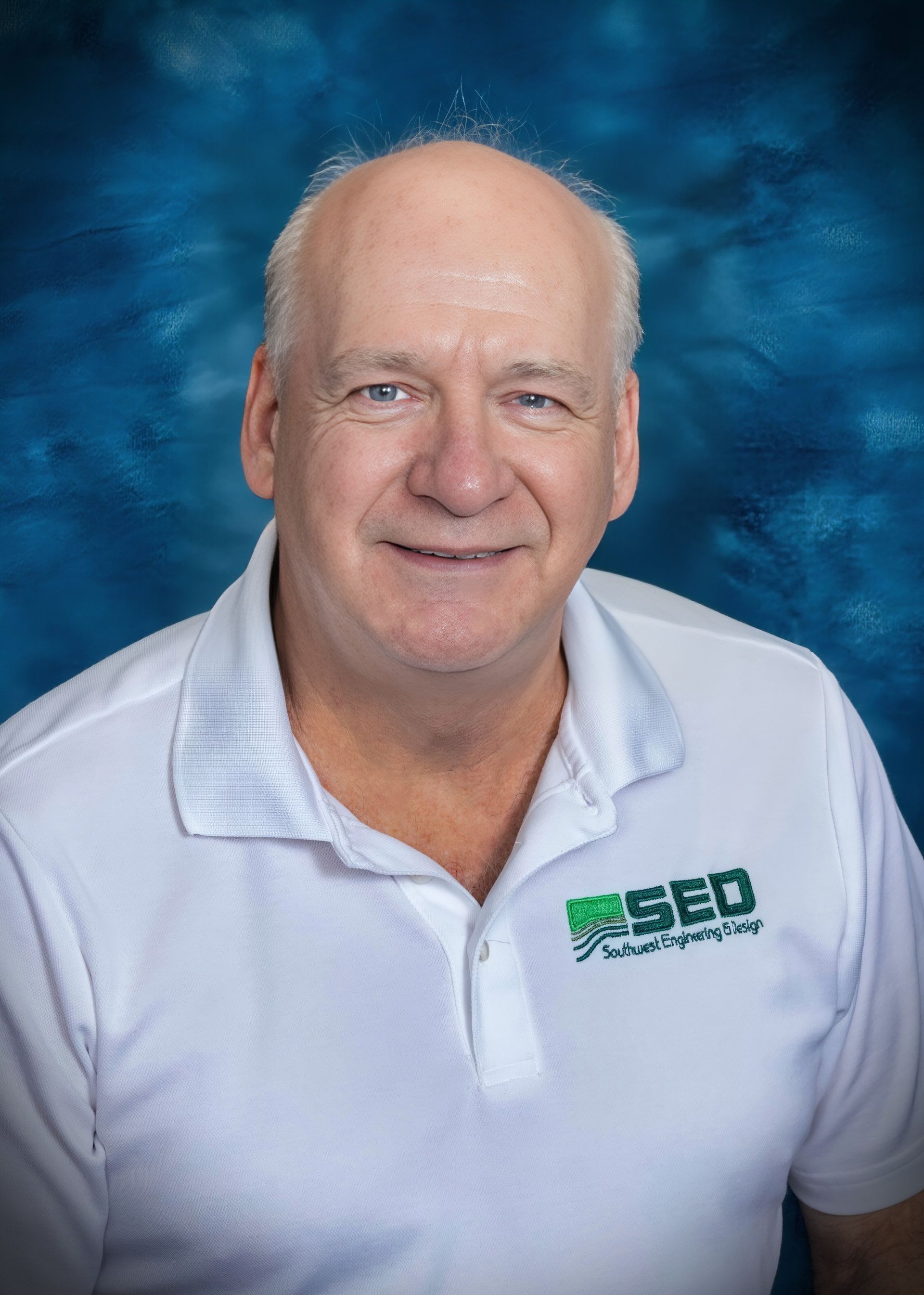 Middle-aged man with fair skin, smiling, wearing a white polo shirt, against a blue background.