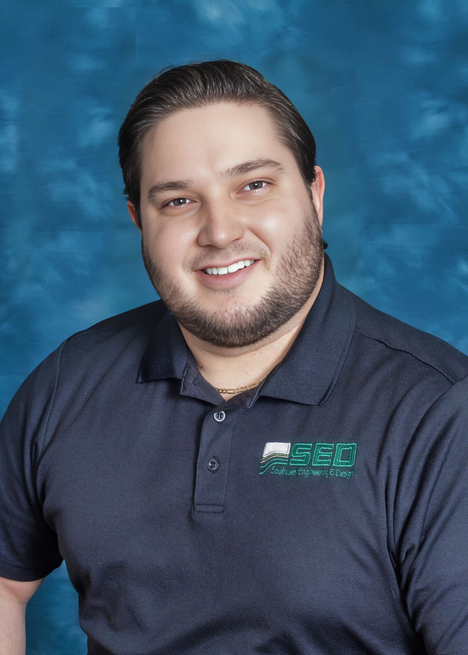Smiling man with dark hair and beard wearing a navy polo shirt, against a blue backdrop.