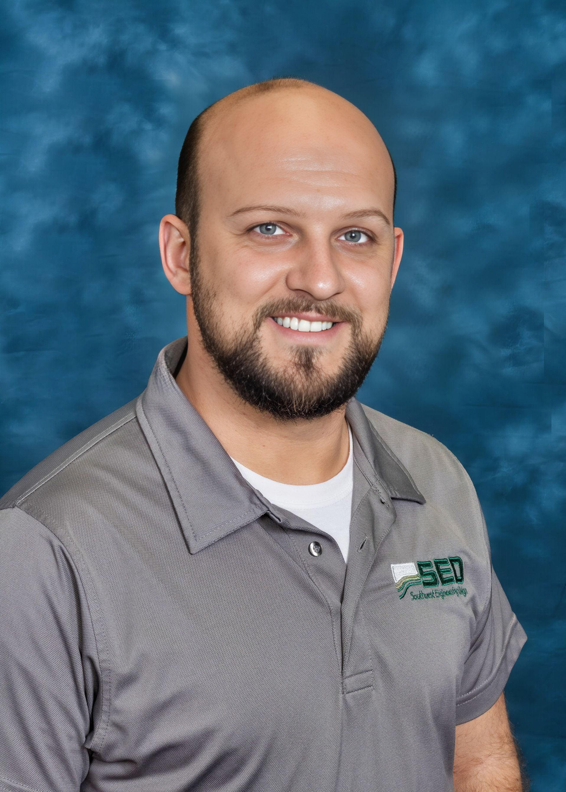 Man with a beard smiling, wearing a gray polo shirt with a logo, against a blue backdrop.